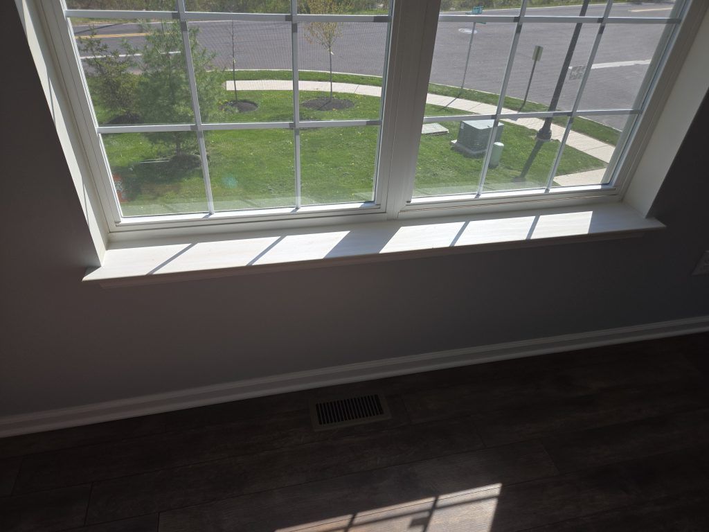 Sunlit empty room with large window, white walls, dark floor, and a view of a sidewalk and grass outside.