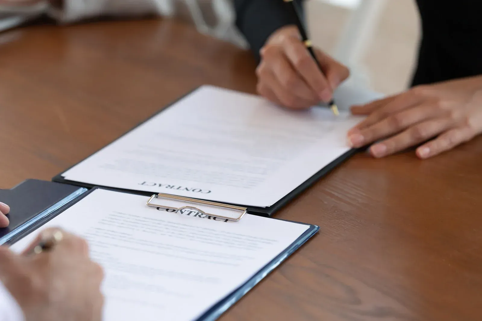 Hands signing documents on a wooden table with clipboards in a meeting room
