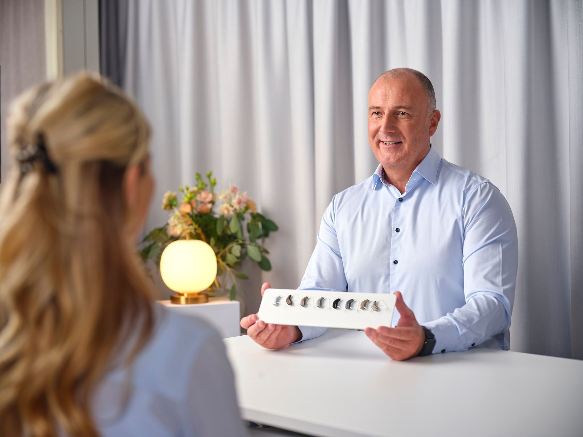 Maður brosir yfir borð, heldur á hvítum spjaldi með litlum táknum í björtu herbergi með blómum og lampa.