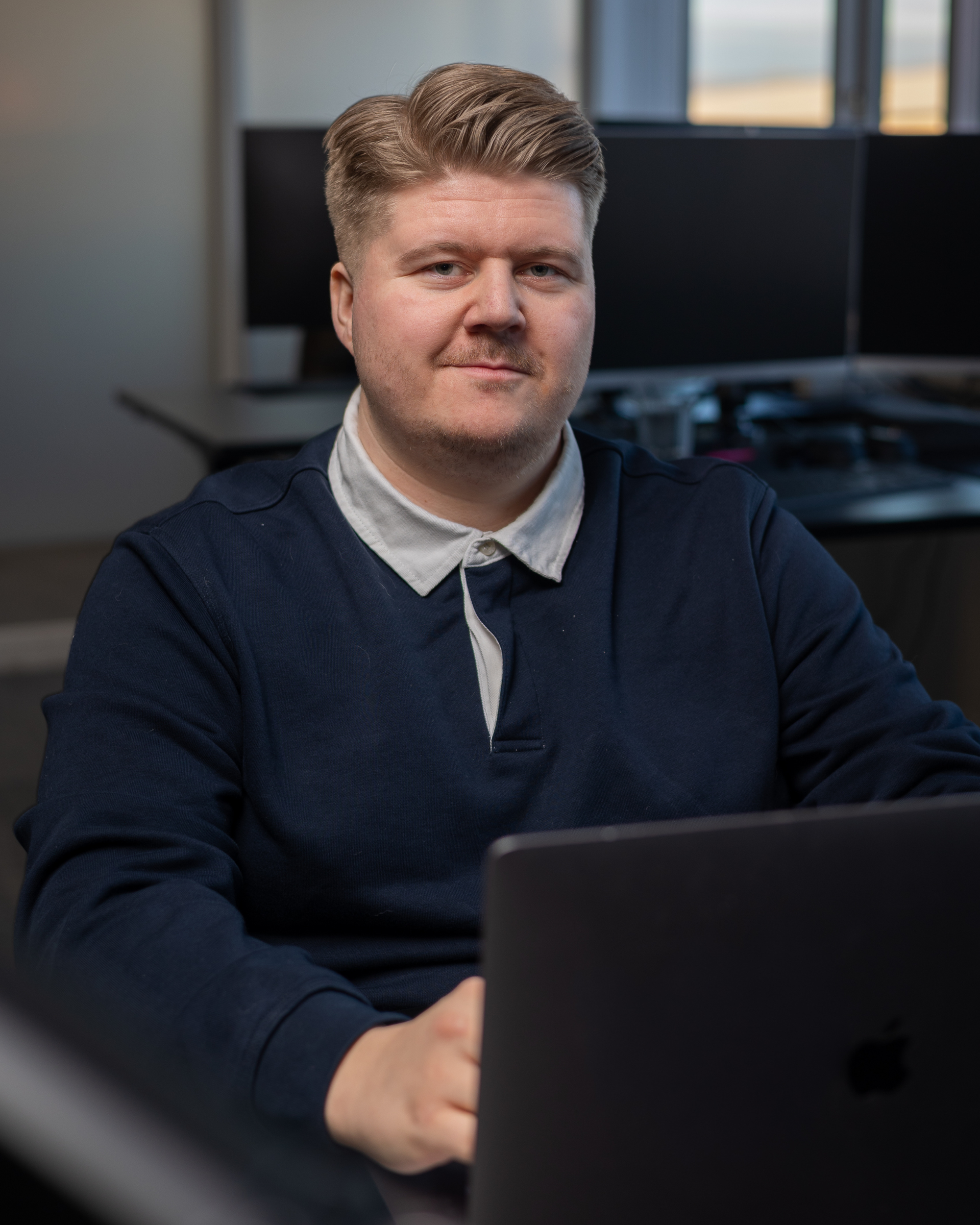 Maður situr við skrifborð með fartölvu á skrifstofu, klæddur í dökkbláa peysu og hvítum skyrtu með kraga.