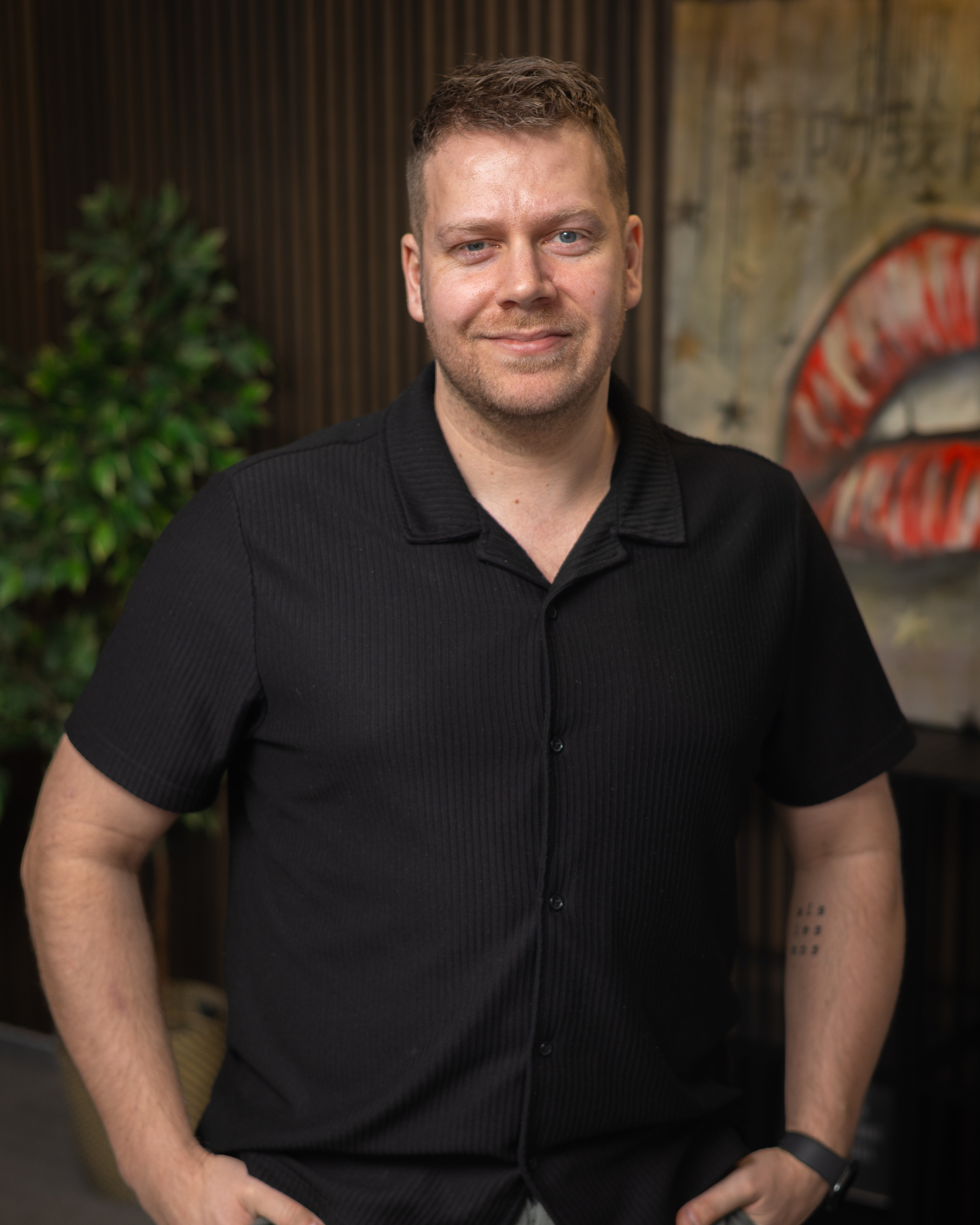 Maður stendur innandyra í svörtum skyrtu, brosandi með hendurnar á mjöðmunum, á sveitalegum bakgrunni.