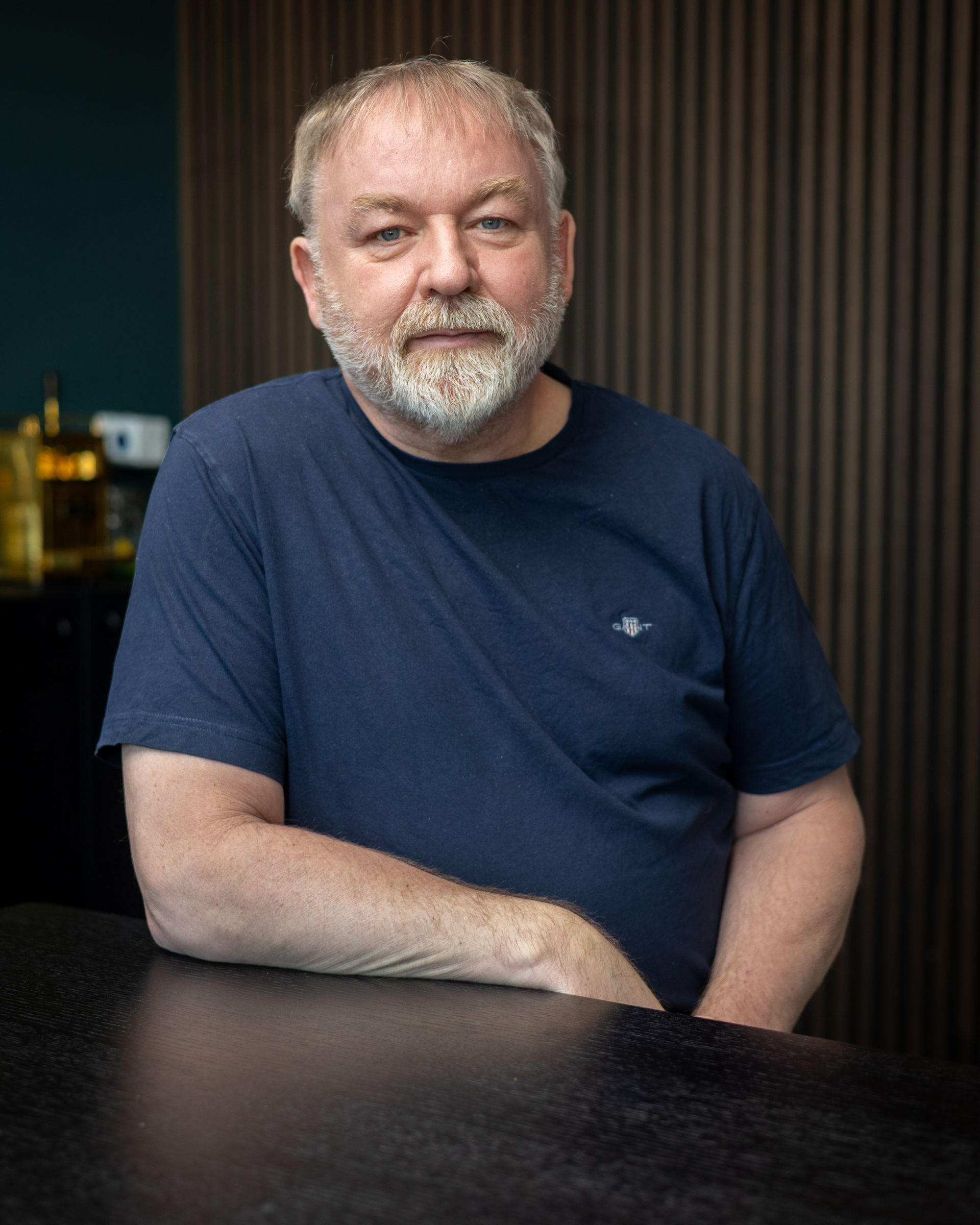 Maður með grátt skegg í dökkbláum bol situr við borð í dimmum herbergi og horfir í myndavélina.