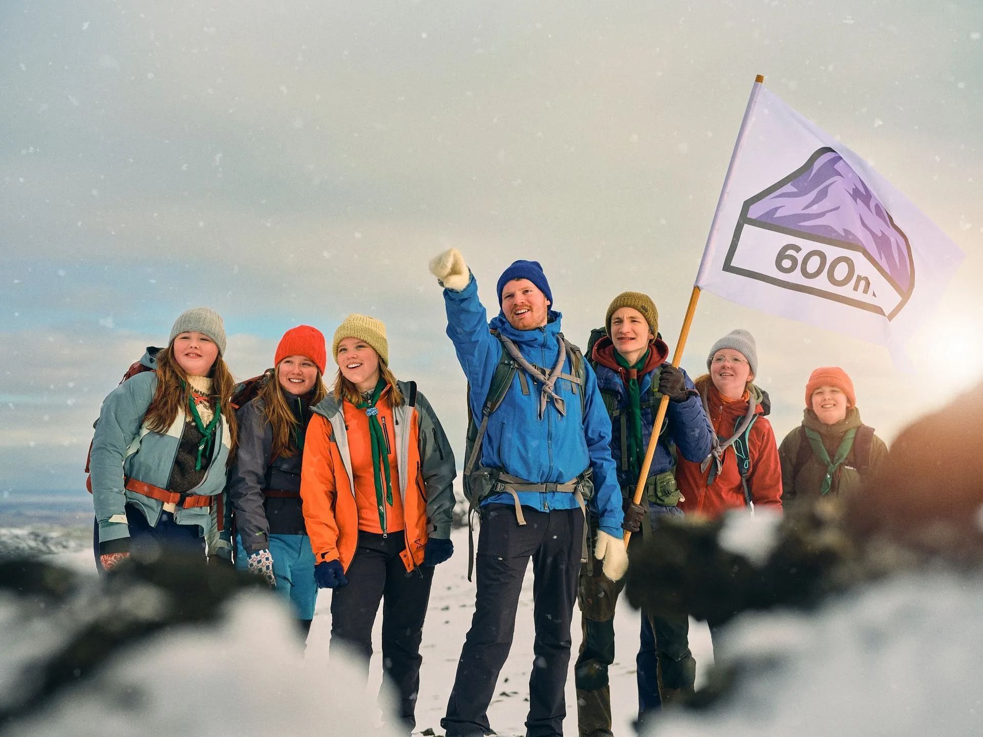 Göngufólk á snæviþöktum hrygg, annar heldur á fjólubláum fána með áletruninni „600“ í köldu fjallalandslagi.