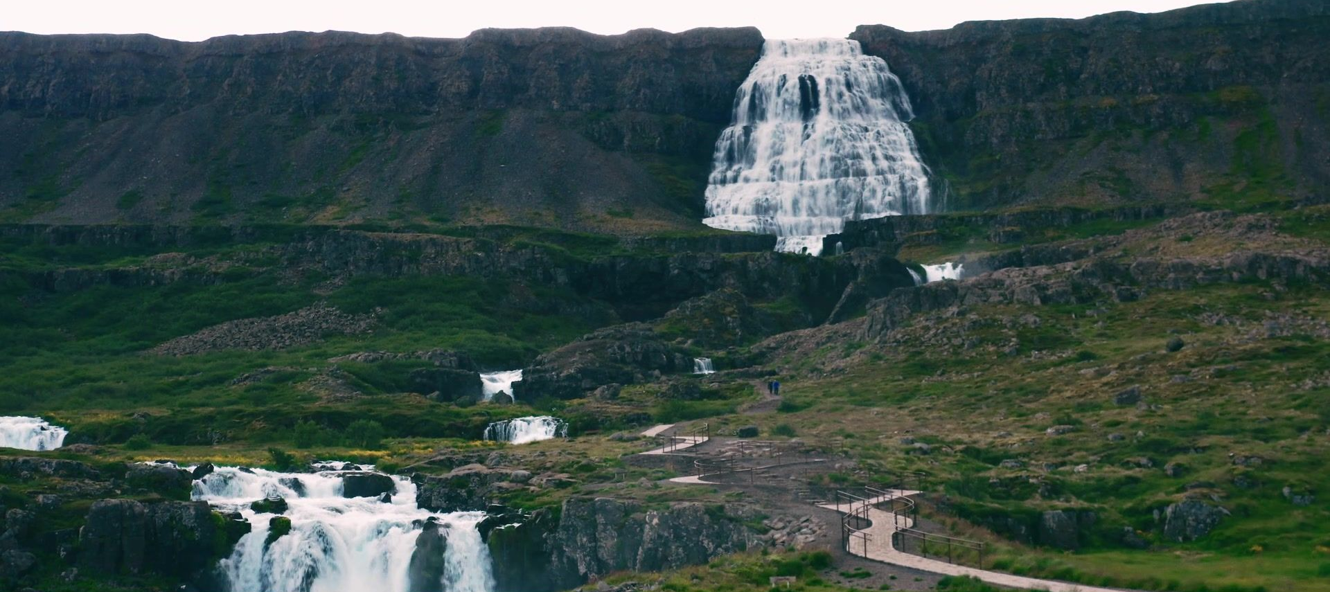 Foss sem fellur niður grænan kletta ofan í gróskumikla dali undir skýjuðum himni