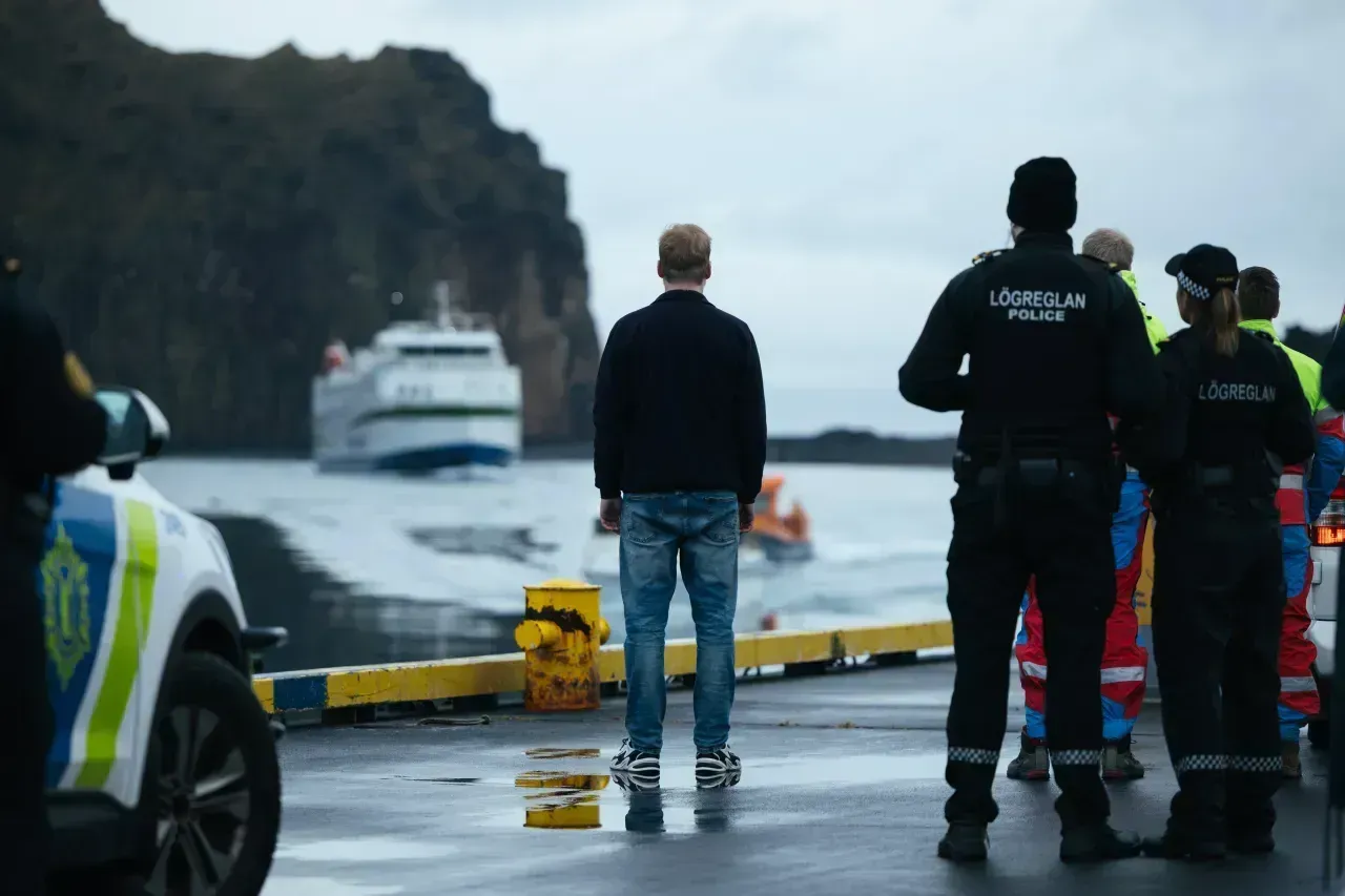 Lögregla og viðbragðsaðilar á blautum hafnarbryggju fylgjast með báti nálægt klettum og appelsínugulum merkimiða.