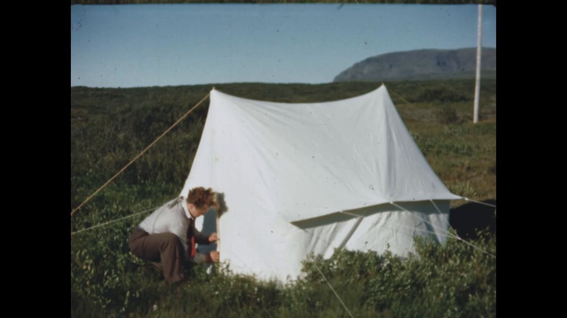 Fólk tjaldar upp hvítum tjaldi á graslendi með hæðum í bakgrunni.