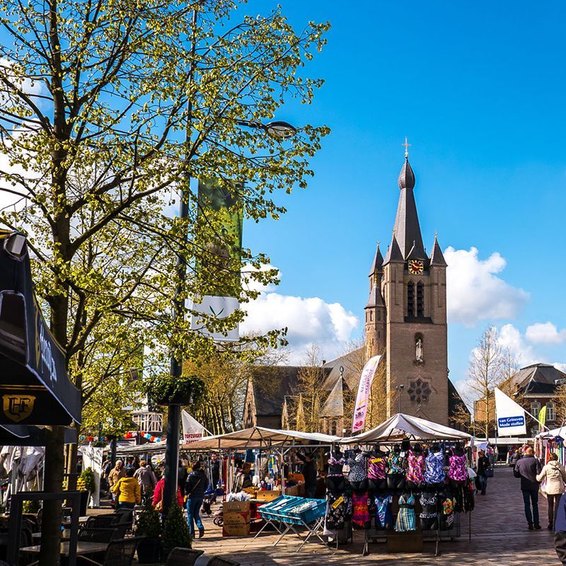 Openluchtmarkt met de kerktoren op de achtergrond. Kopers struinen langs de kraampjes onder een strakblauwe hemel.