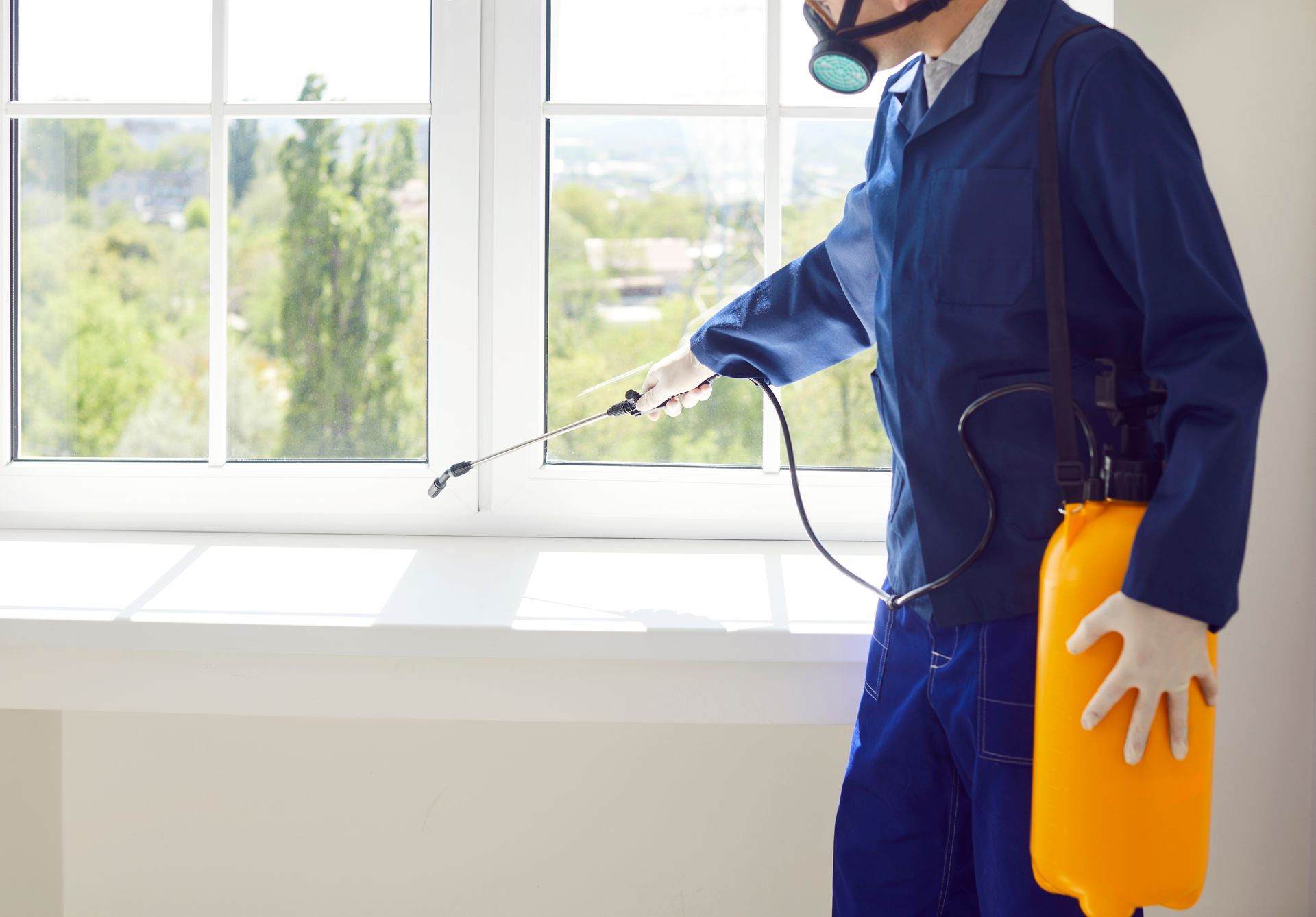 Technician in protective gear spraying pesticide along a windowsill inside a commercial building. Pest control worker in blue coveralls spraying pesticide near a window, holding a yellow container