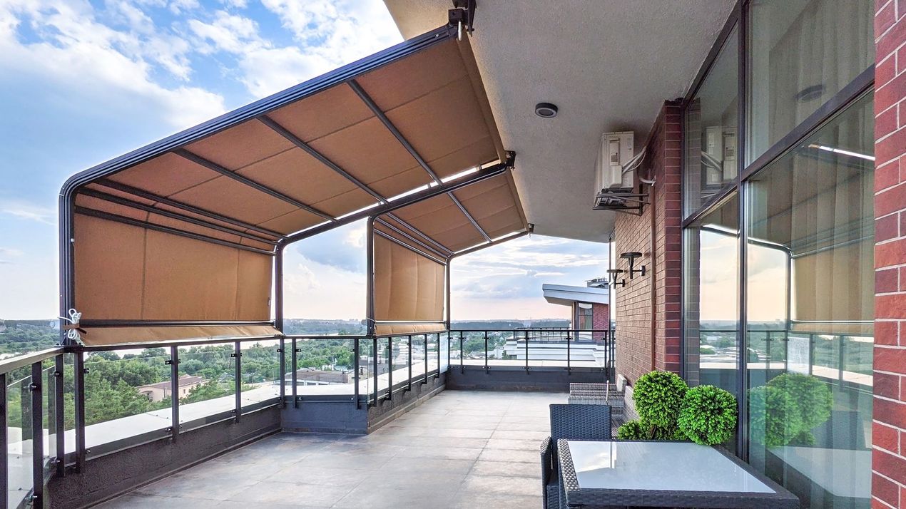 Una tenda da sole retrattile color beige si estende su un balcone con ringhiere in vetro, un tavolino e piante da esterno.