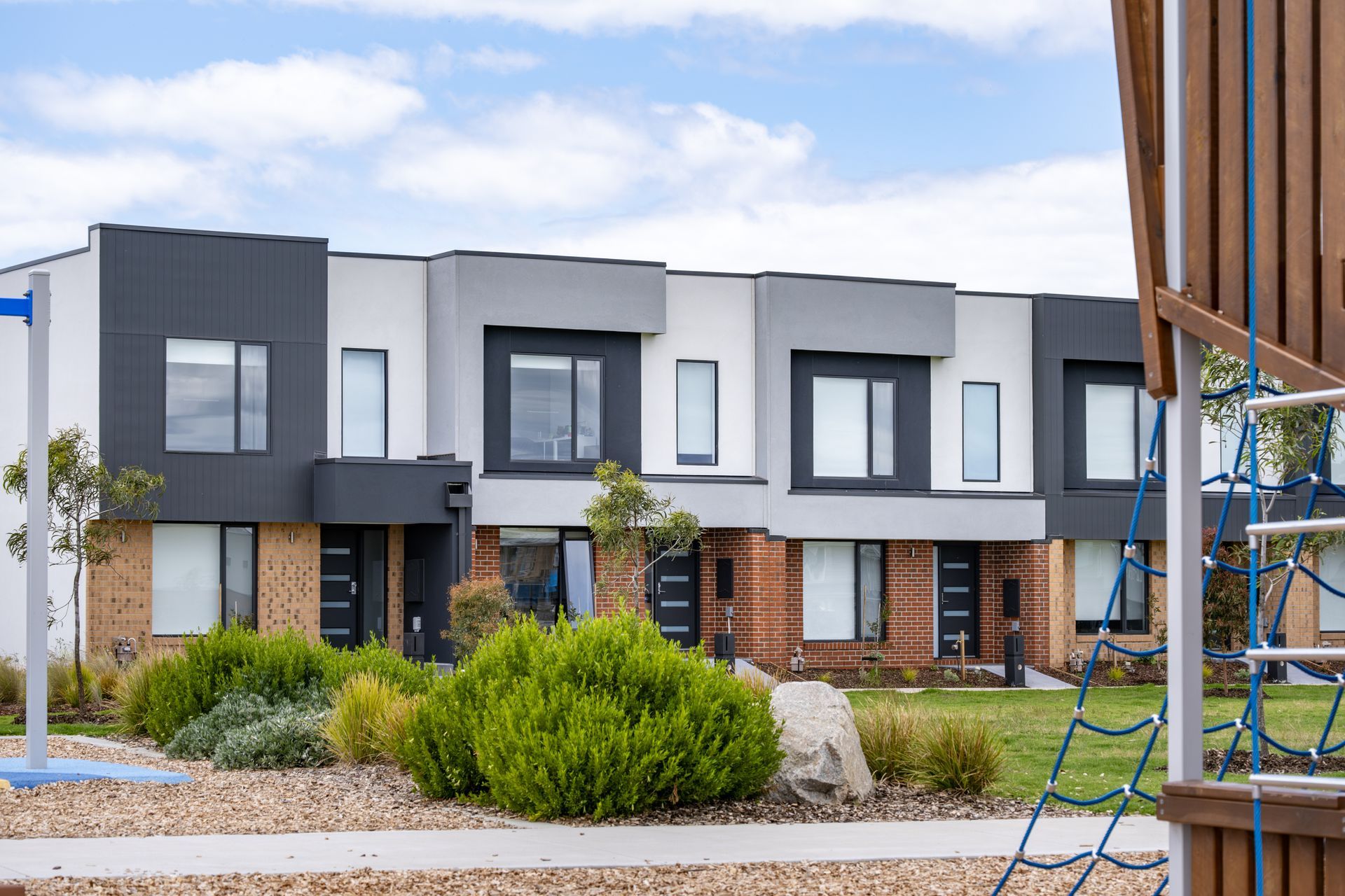 Row of modern townhouses with gray, white, and brick exteriors; shrubs and playground in foreground.