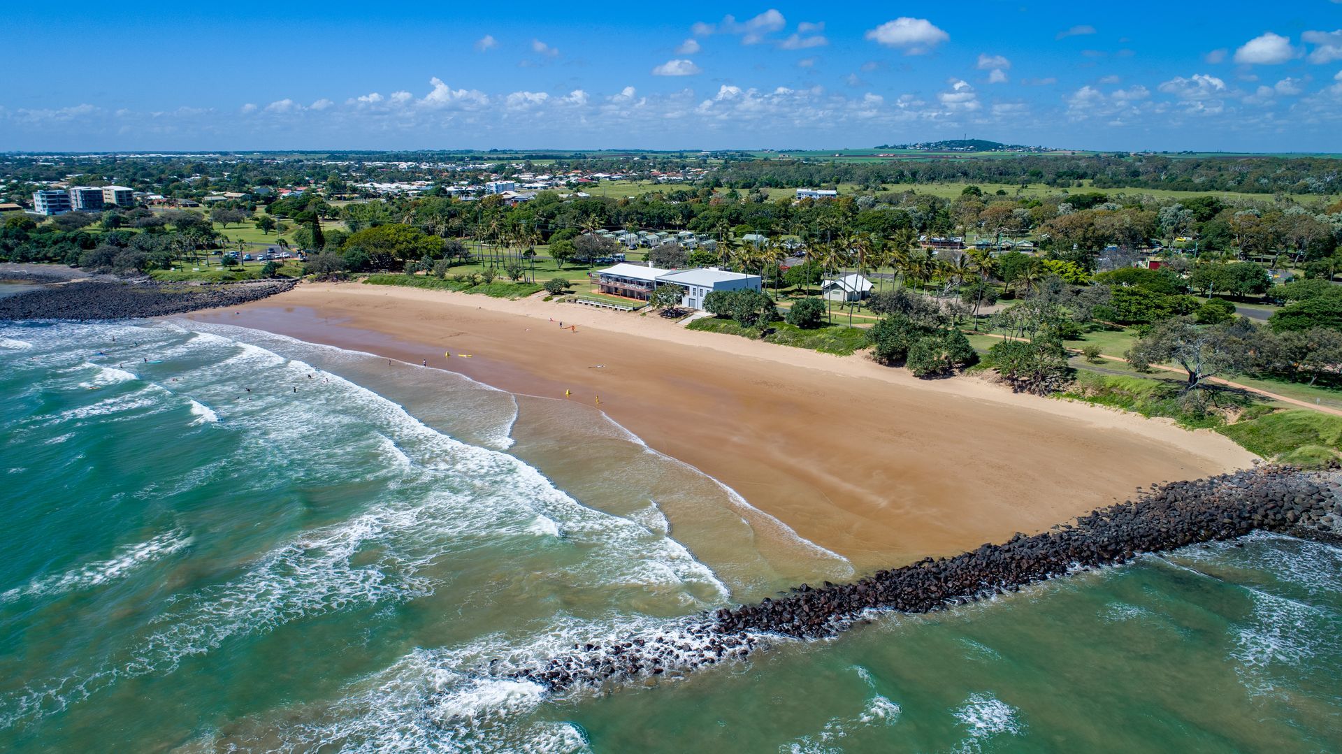Aerial view of a sandy beach with turquoise water, a rocky breakwater, and buildings along the shore. Aerial view of a sandy beach with turquoise water, a rocky breakwater, and buildings along the shore.