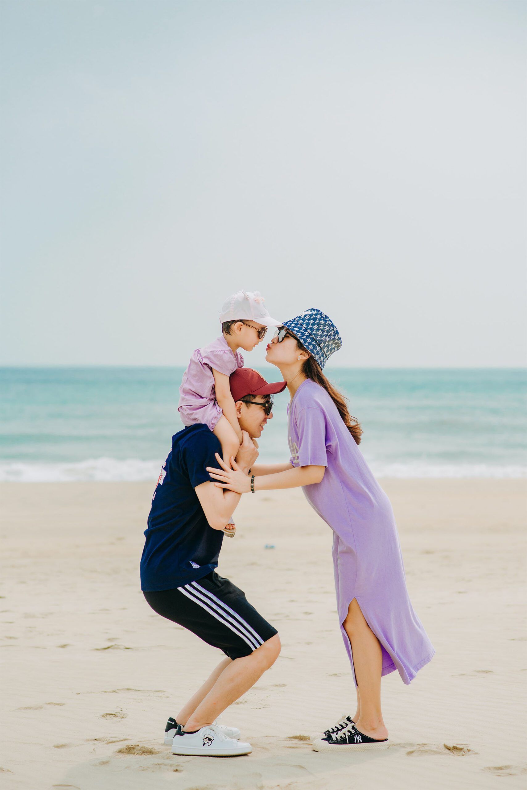 Family on beach: Dad carrying child on shoulders, mom kissing, ocean in background.