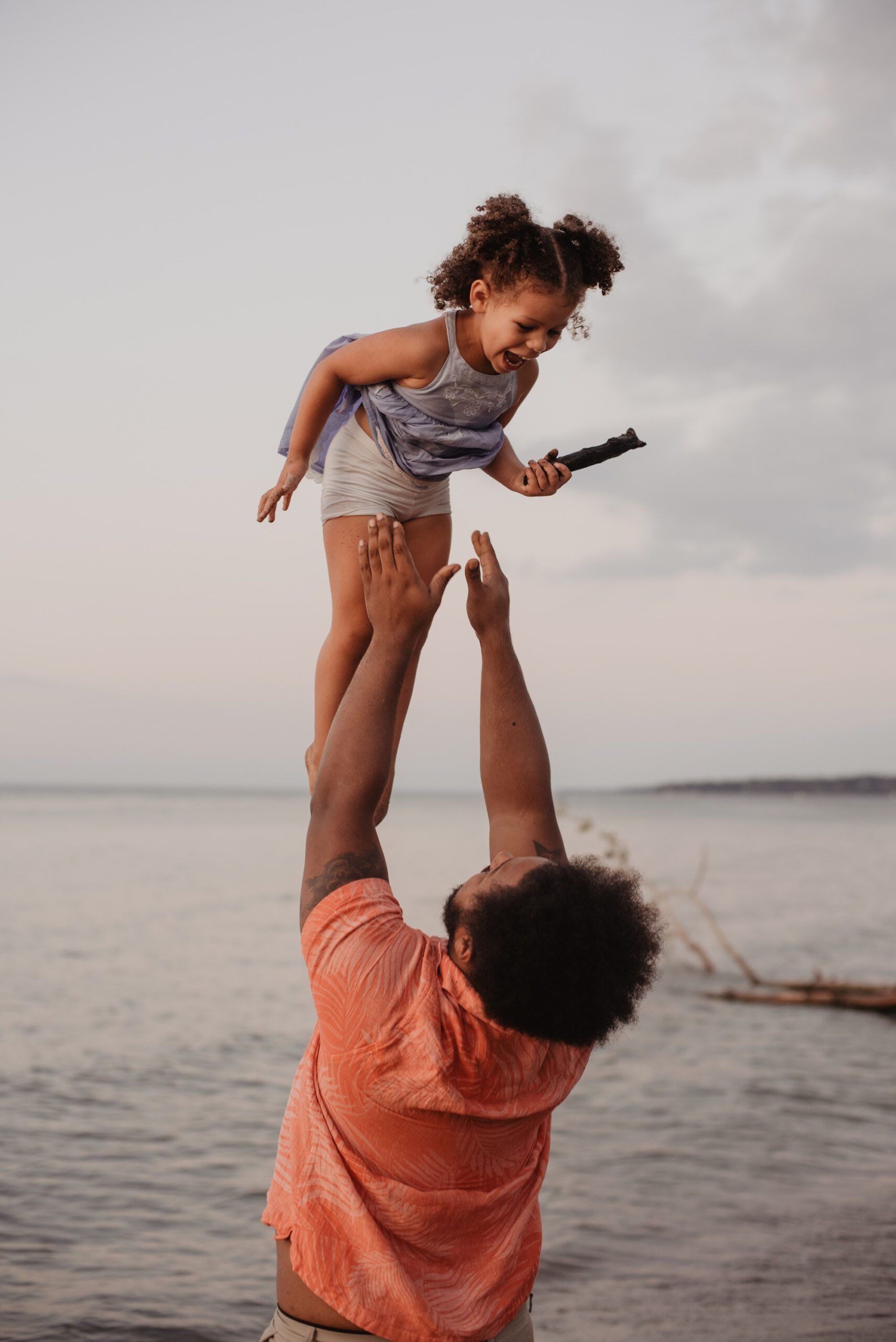 Father lifts a child into the air near a body of water. Child holds a dark object and smiles.