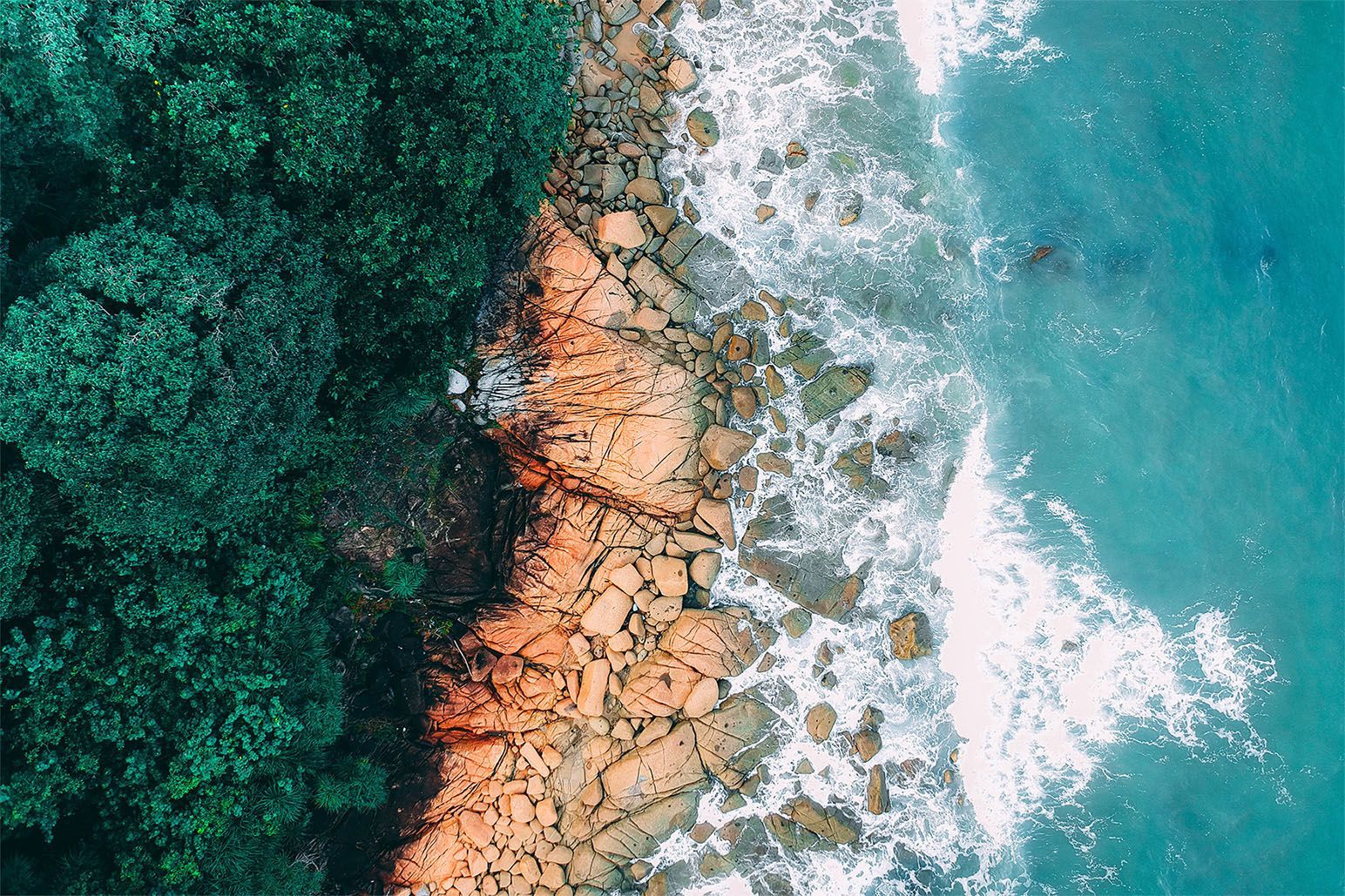 Aerial view: Ocean waves crashing on rocky shoreline next to lush green forest.