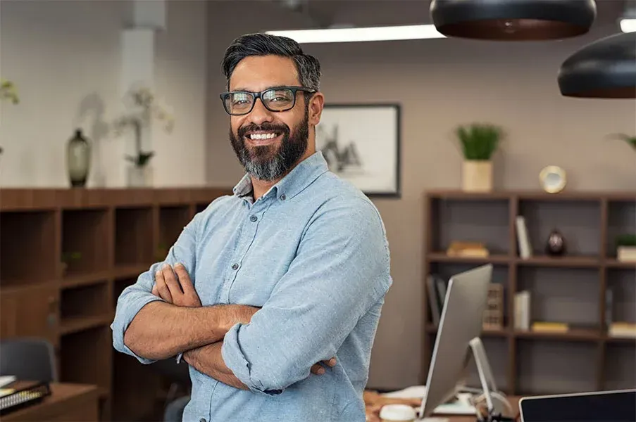 Man with glasses smiles, arms crossed, in an office.