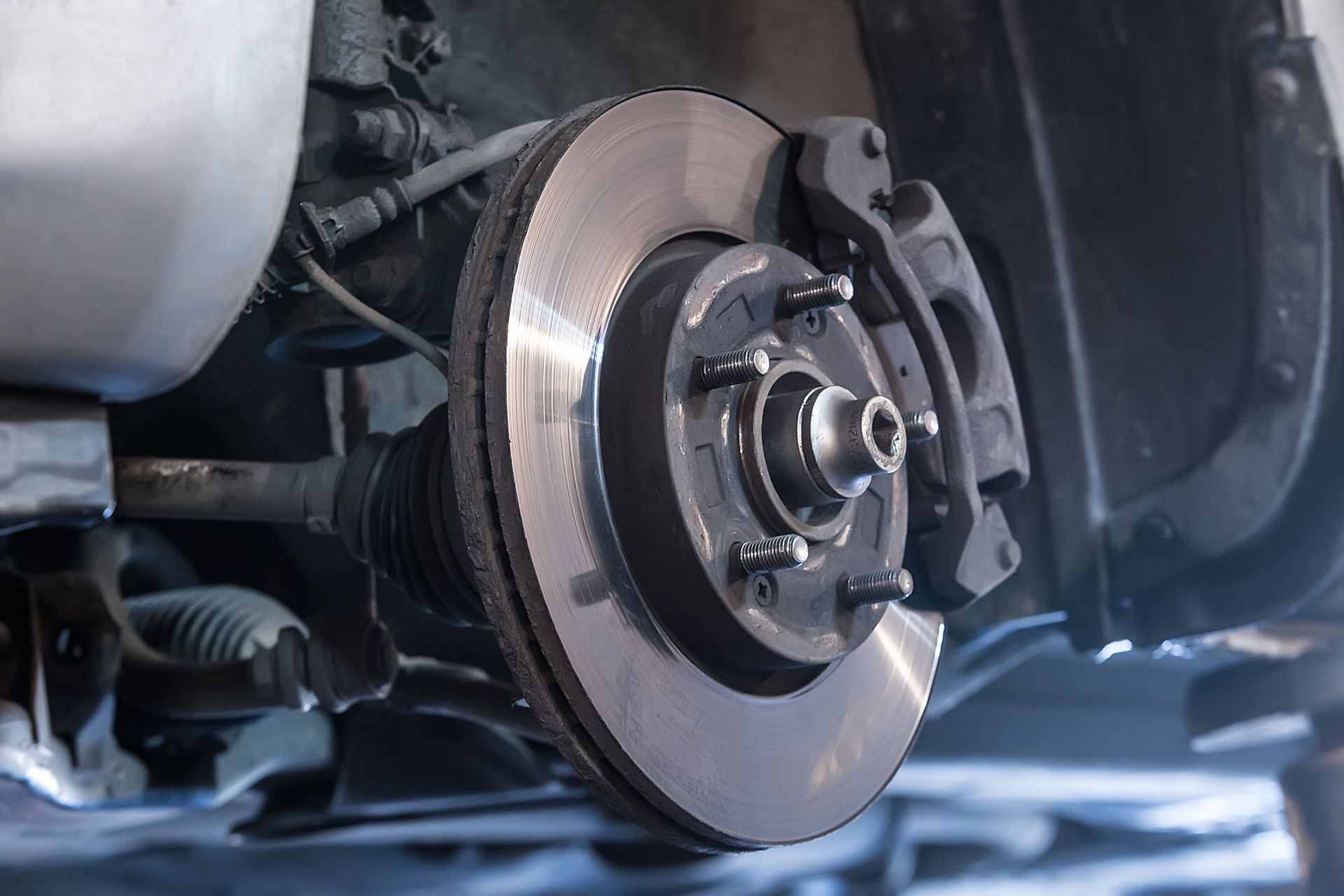 Close-up of a vehicle’s brake disc, caliper, and suspension components viewed from underneath the chassis.