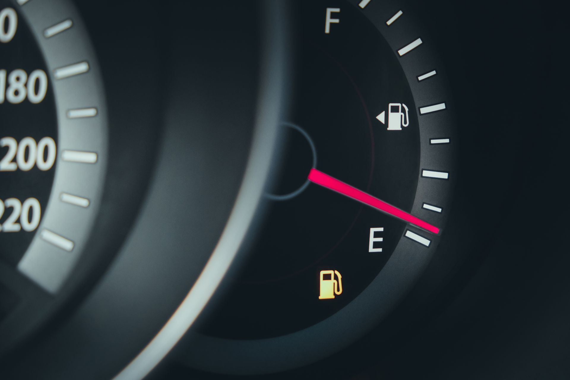A close-up of a car fuel gauge with the needle pointing near E and a yellow low-fuel warning light illuminated.