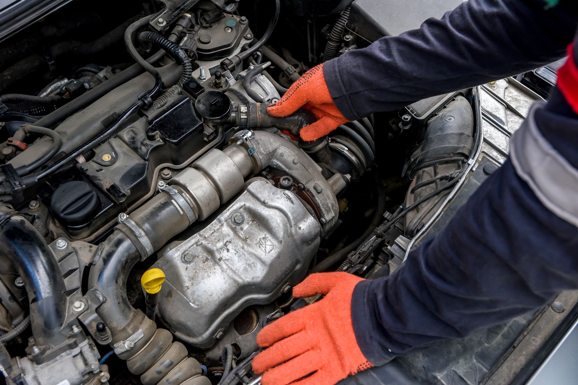 A mechanic wearing orange work gloves inspects the engine of a car.