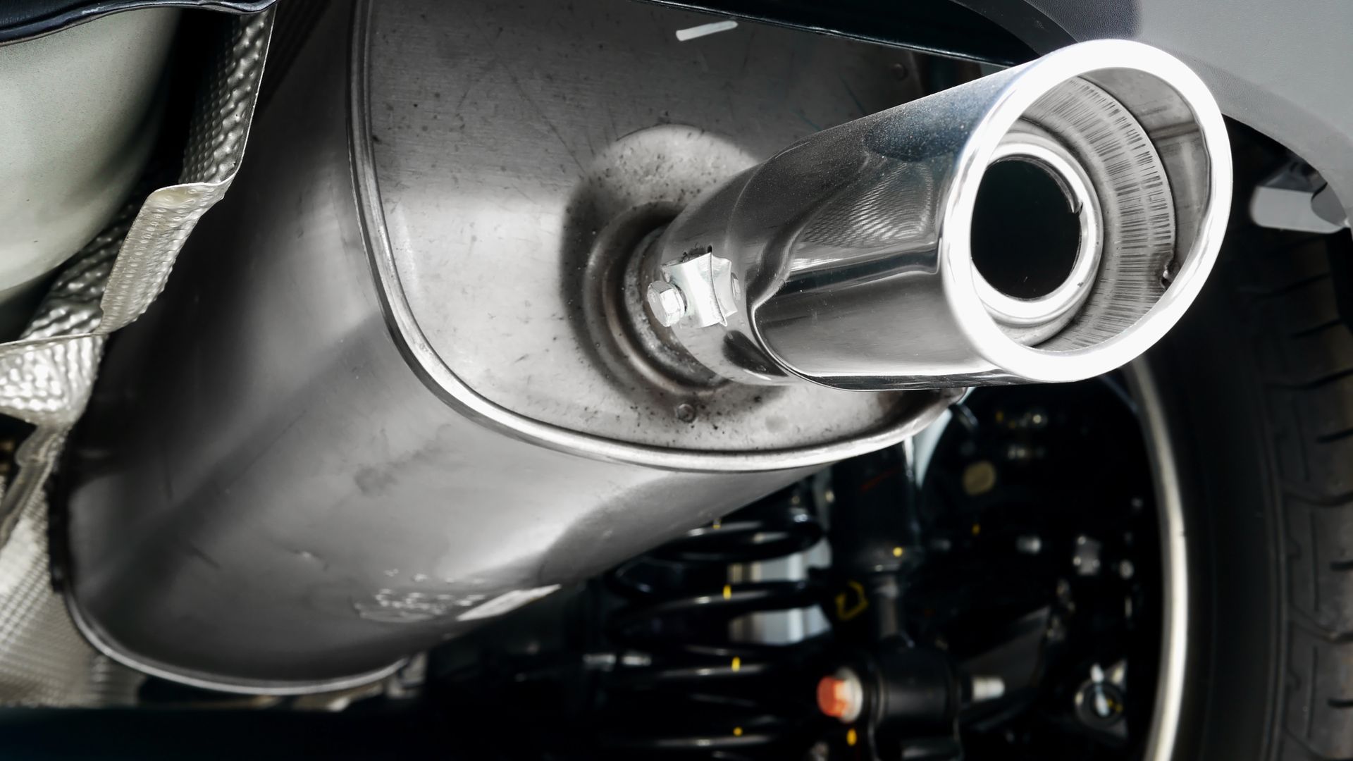 A close-up, underside view of a clean, metallic car muffler and exhaust pipe extension, with a suspension spring visible.