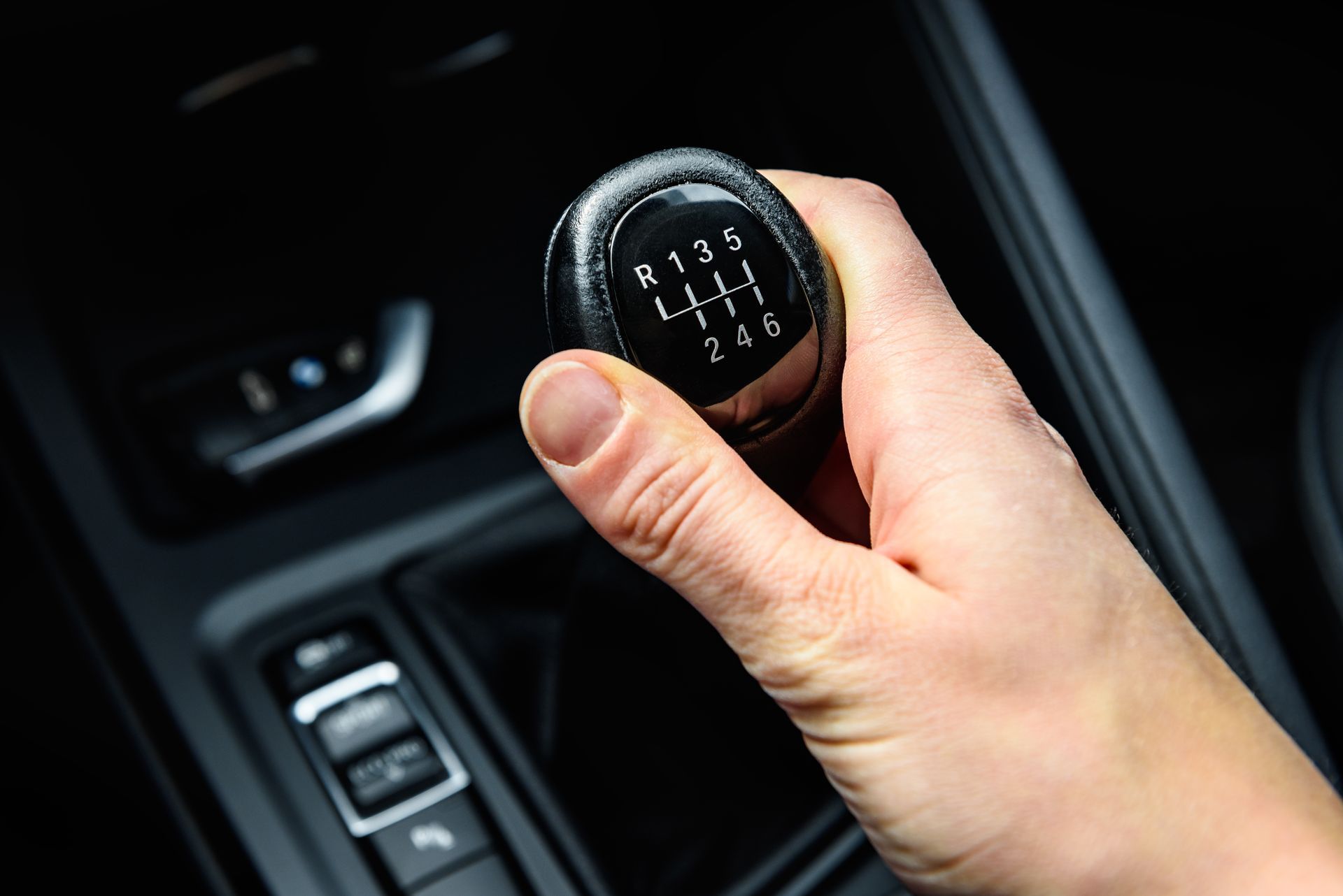 A hand gripping a manual car gear shift knob, showing a six-speed transmission pattern in a modern vehicle interior.