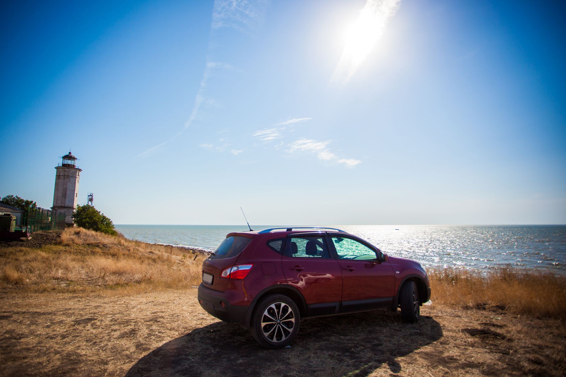 A dark red crossover SUV parked on a grassy cliffside overlooking the sea near a white lighthouse under a sunny blue sky.