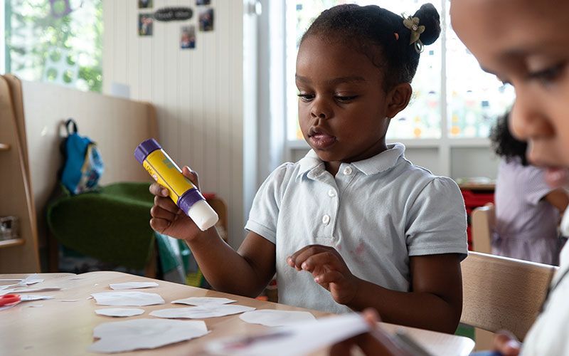 A young girl is sitting at a table holding a glue stick.