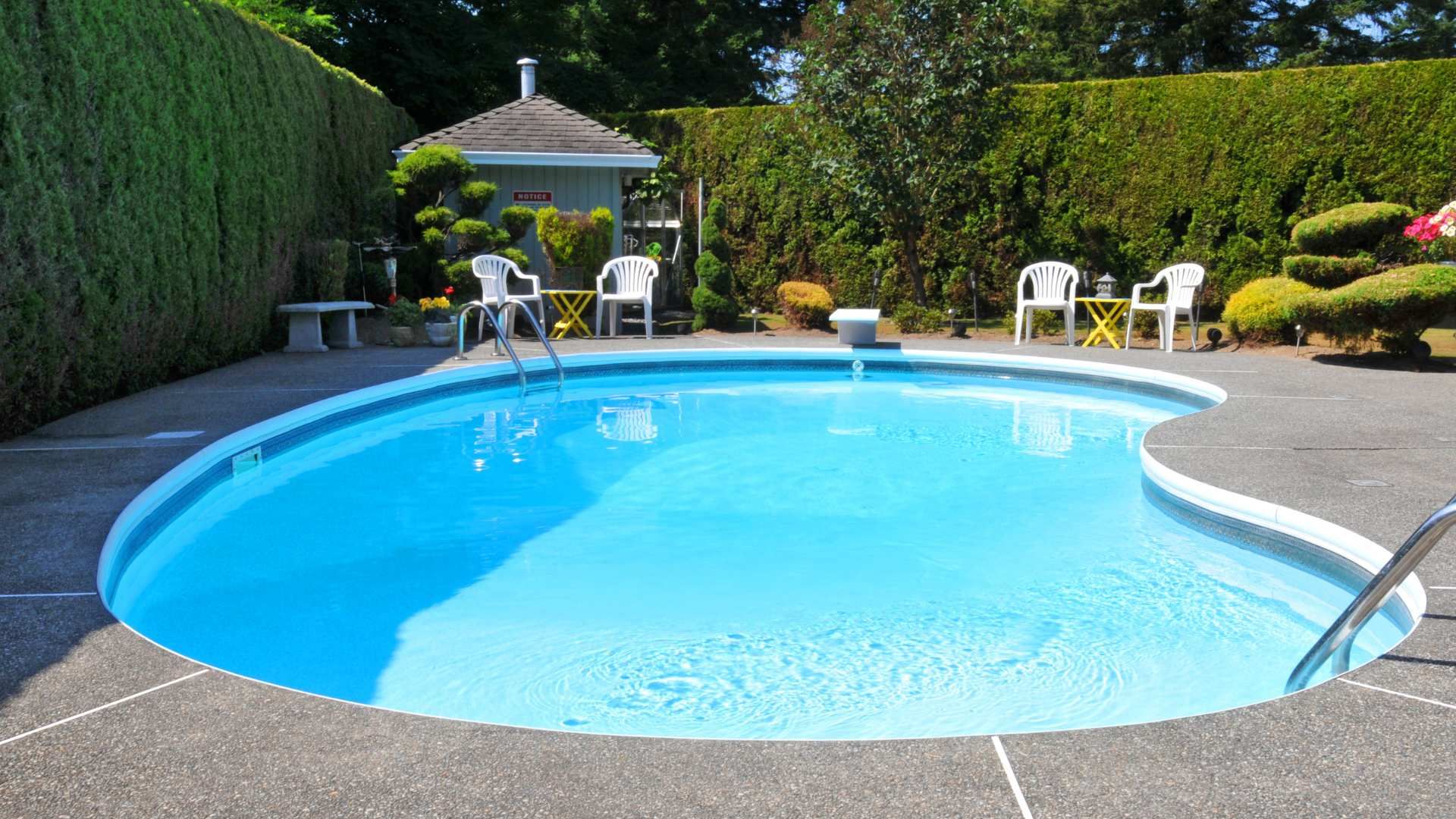 Backyard swimming pool with blue water, patio chairs, and a white gazebo surrounded by hedges.