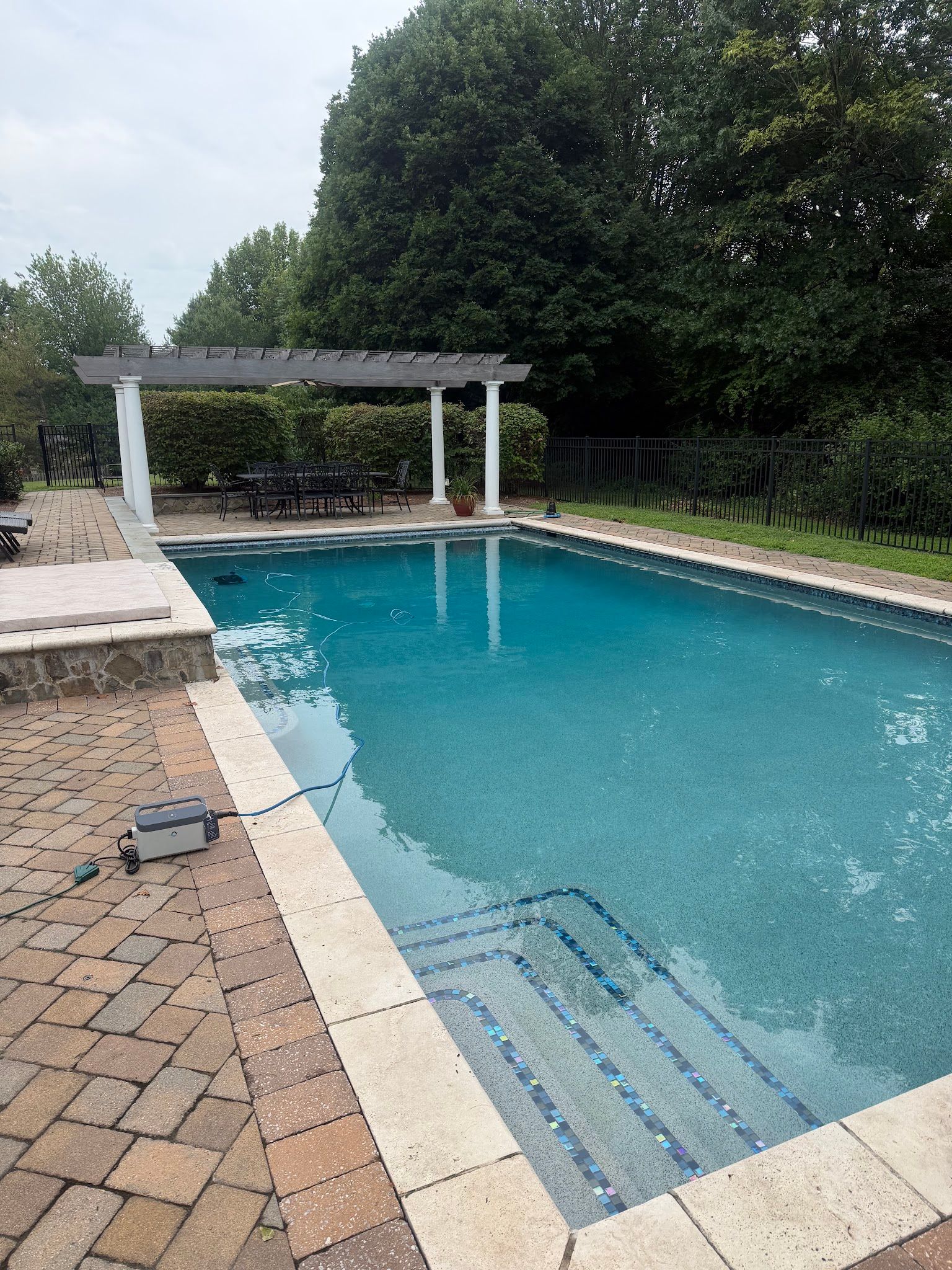 Outdoor swimming pool with blue water, brick deck, and white shade structure beside trees