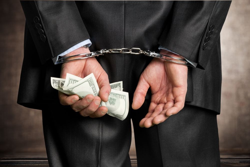 A man in a suit and handcuffs is holding a bunch of money behind his back.