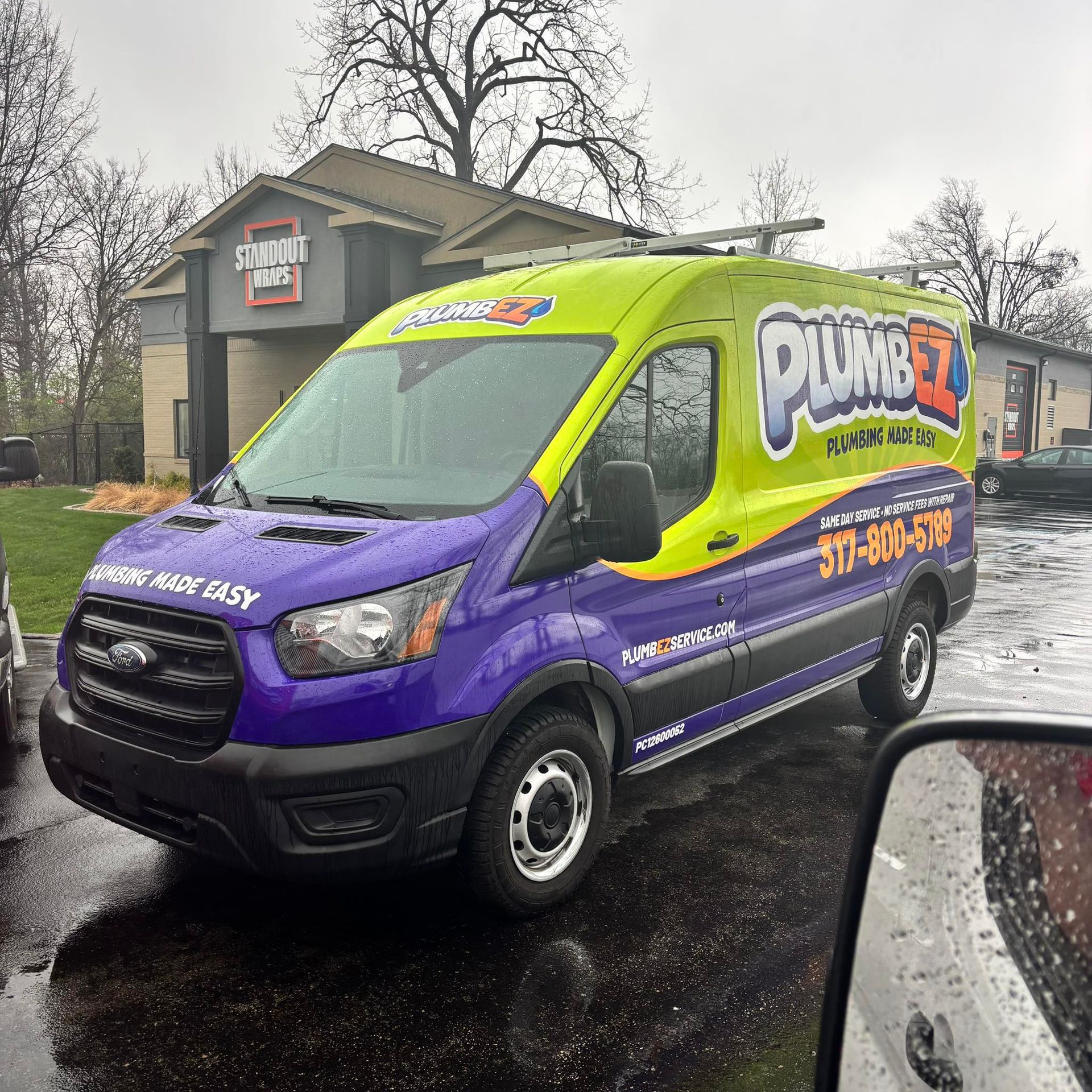 Bright purple and lime green plumbing service van parked on a wet street in front of a house.