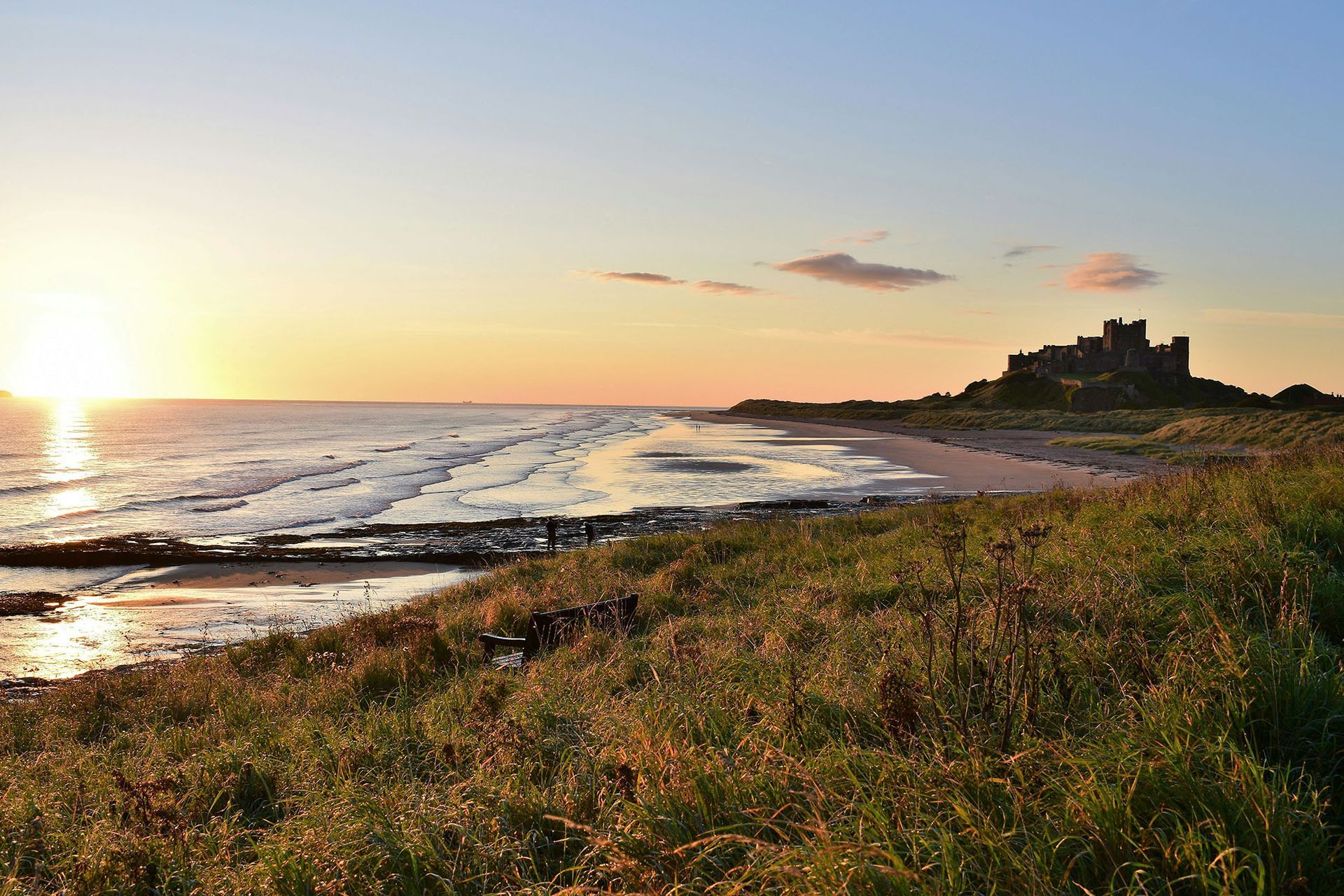 Sunset over a rocky coastline with waves, grassy foreground, and a silhouetted hilltop castle