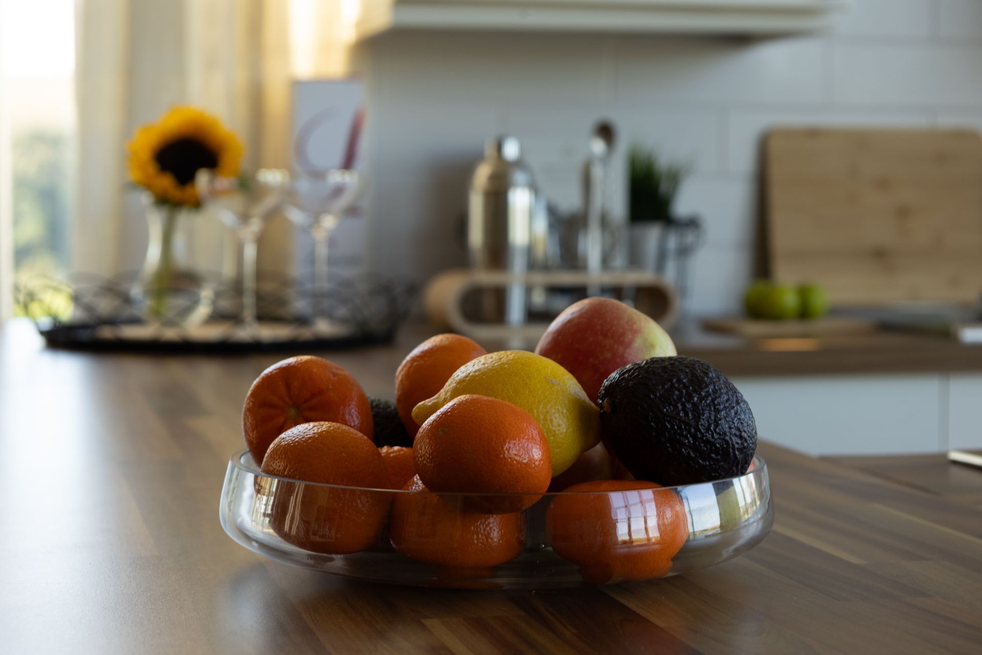 A glass bowl of assorted fruit on a kitchen counter with a blurred sunflower in the background