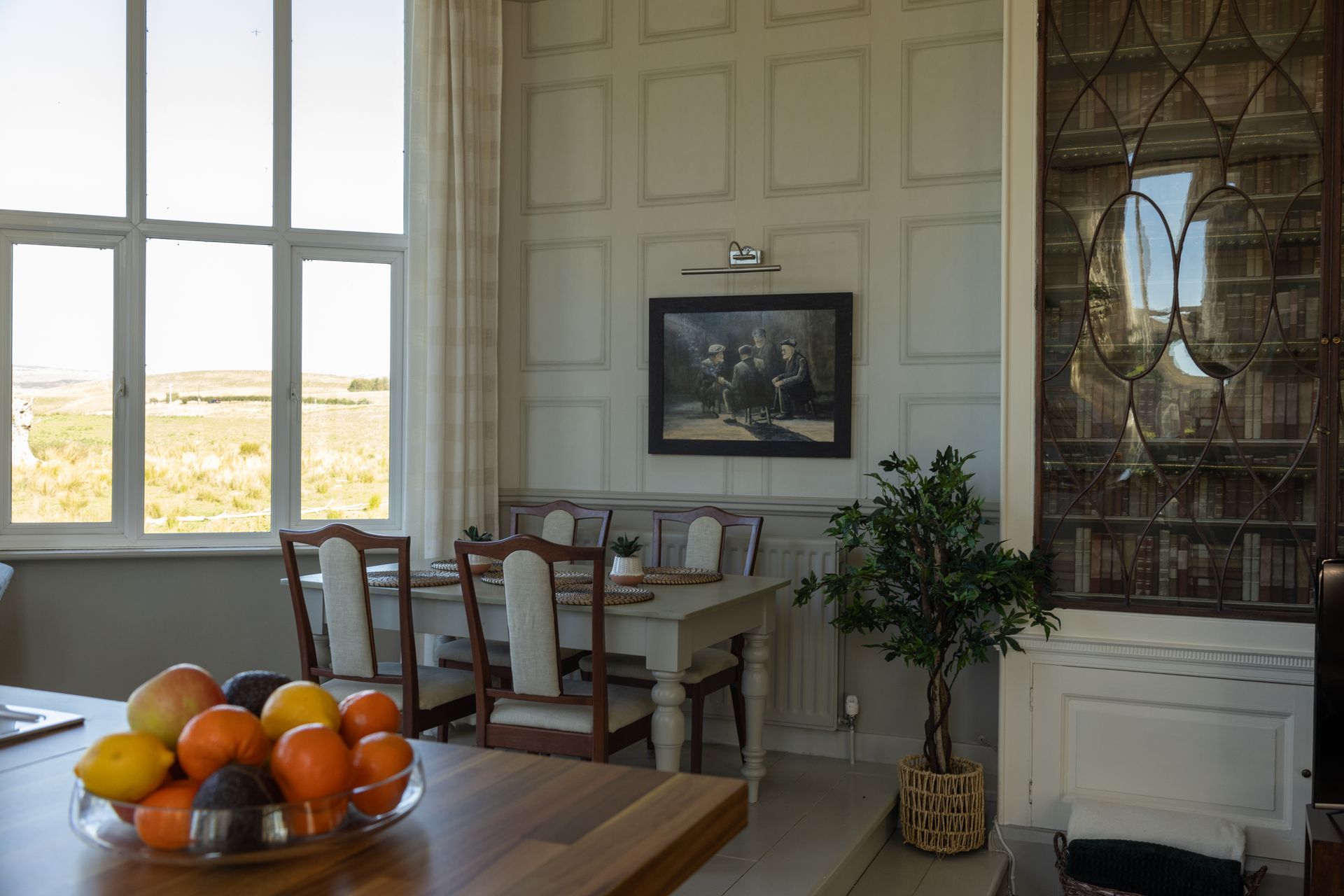 Bright room with table of fruit, chairs, white cabinet, framed art, and windowed view outside