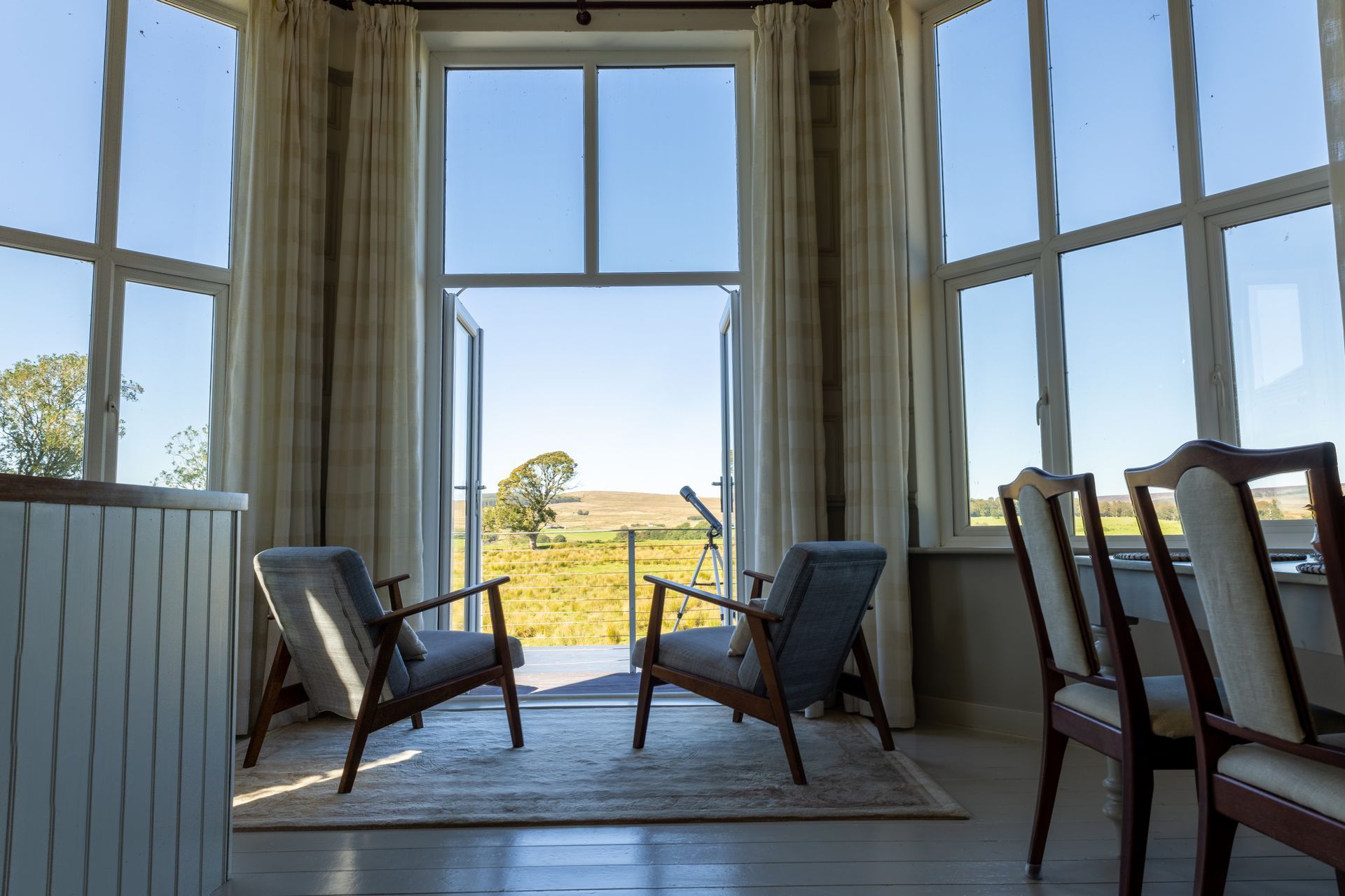 Two lounge chairs by tall windows overlooking a sunny countryside view