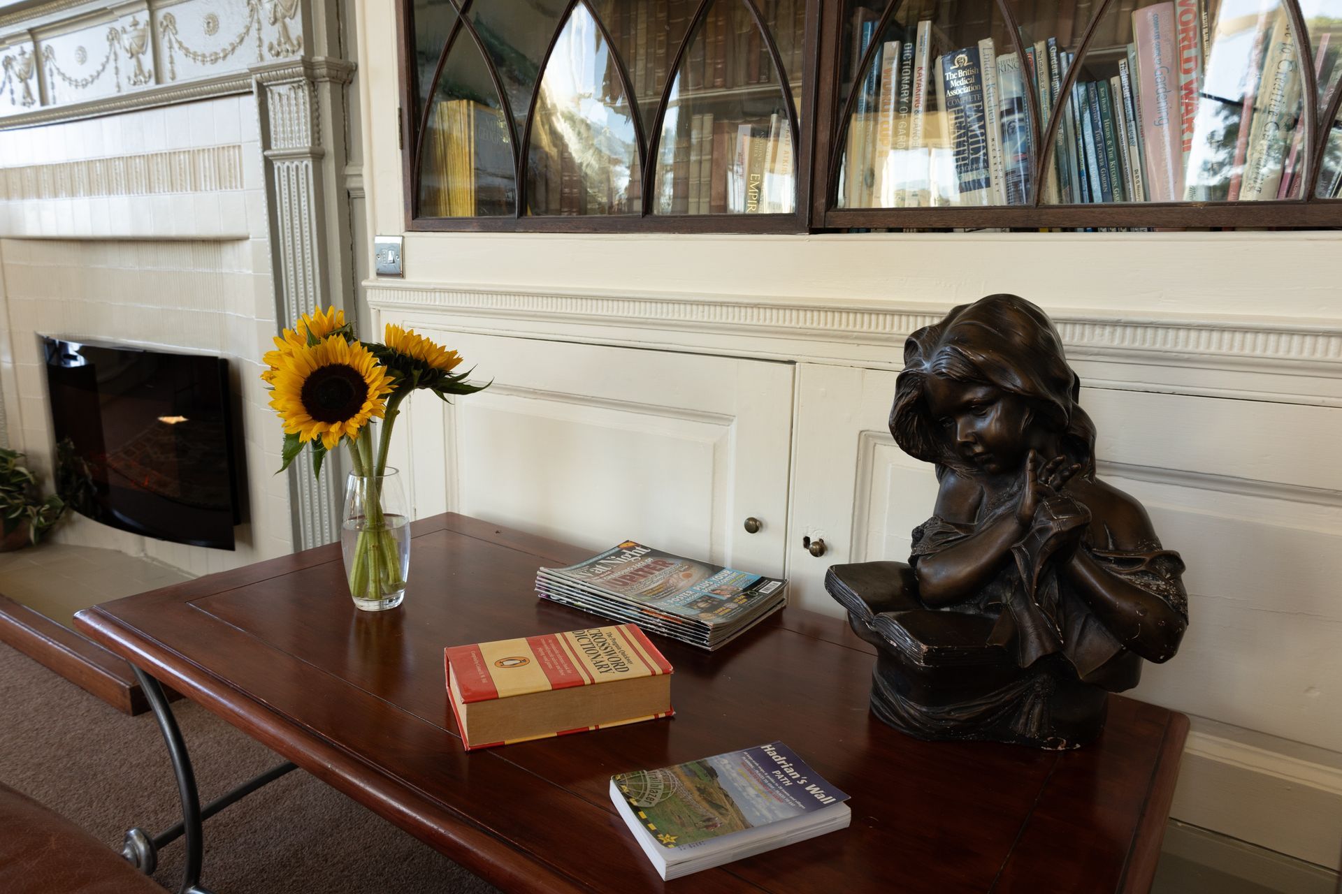 Dark wood table with books, a bust, and sunflowers beside a fireplace in a formal room