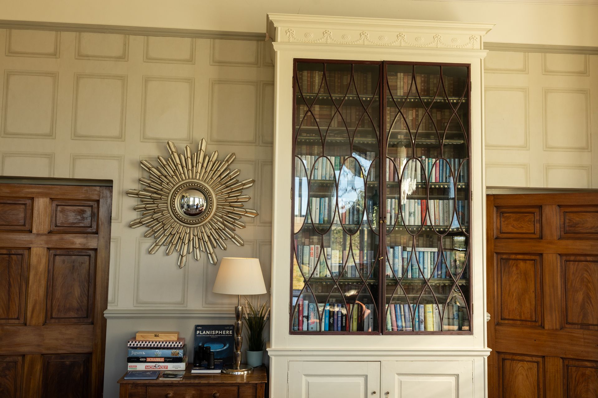 Living room wall with a tall mirrored cabinet, sunburst mirror, lamp, books, and wood-paneled doors