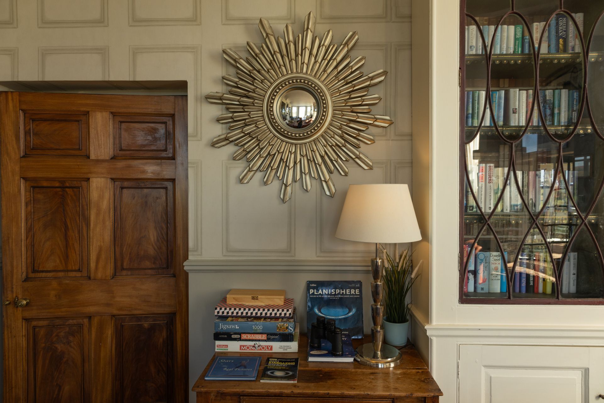 Elegant room corner with paneled wood door, sunburst mirror, lamp, books, and glass-front shelving