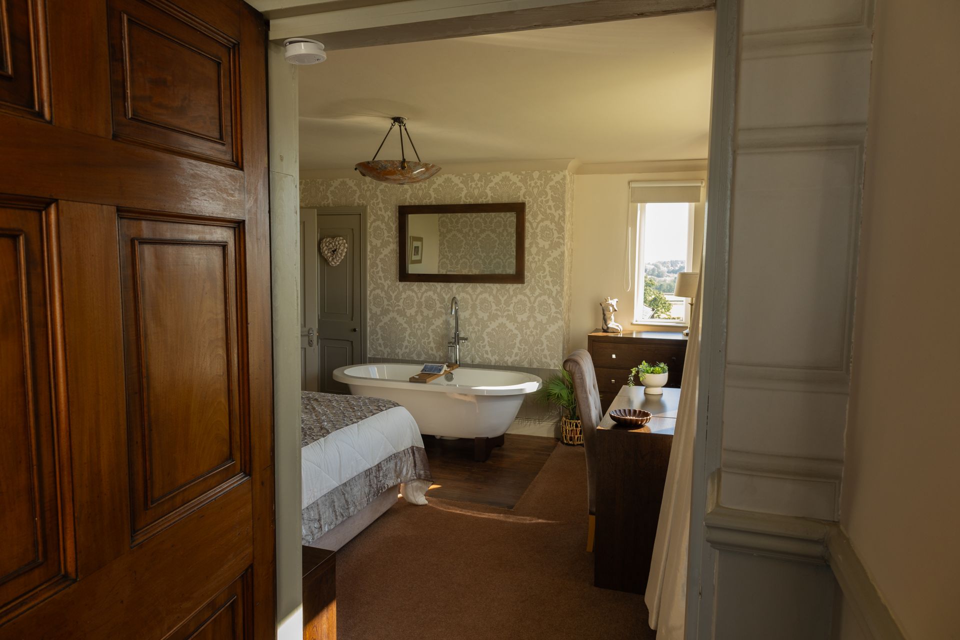 View through a doorway into a bright bedroom with a bed, bathtub, desk, and window.
