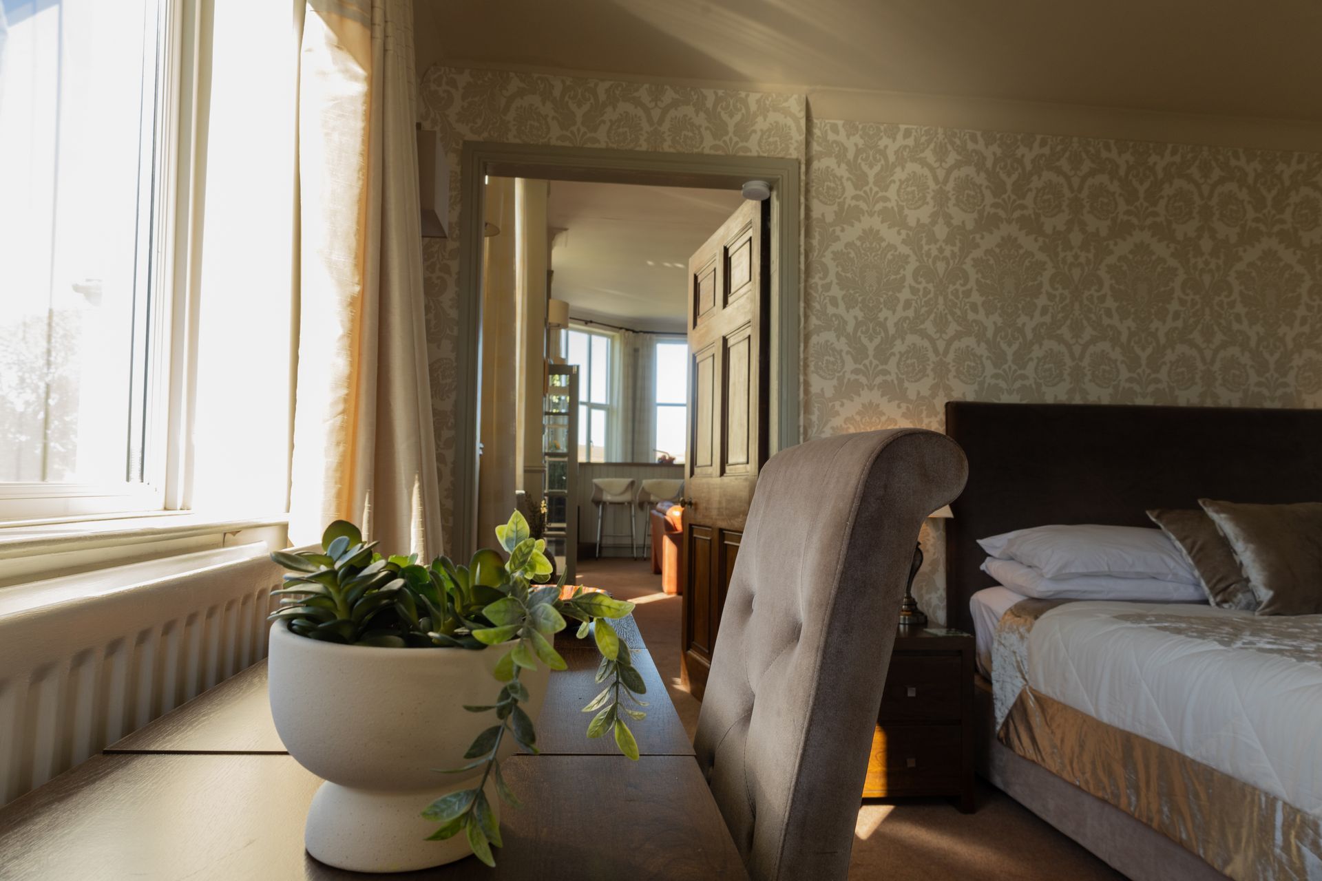 Sunlit bedroom with bed, potted plant, patterned wallpaper, and window with sheer curtains