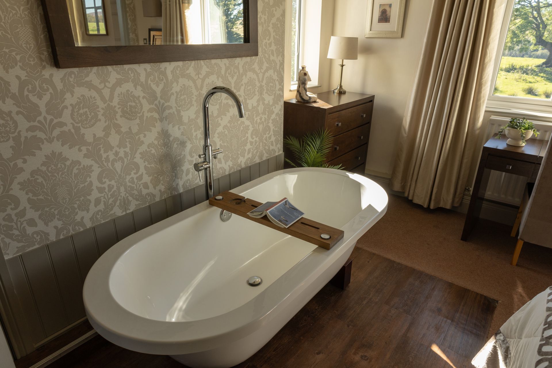 Bathroom with freestanding white tub, wooden bath tray, and window with curtains and garden view