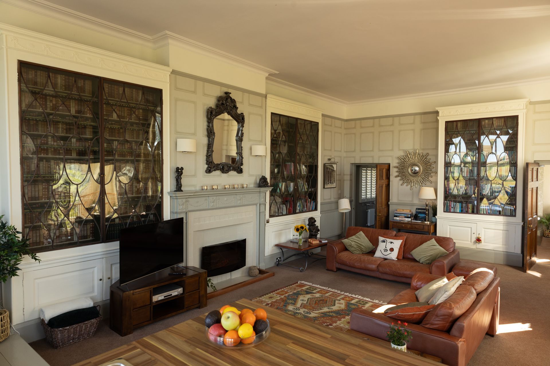 Cozy living room with leather sofas, fireplace, patterned rugs, and tall windows with dark drapes.