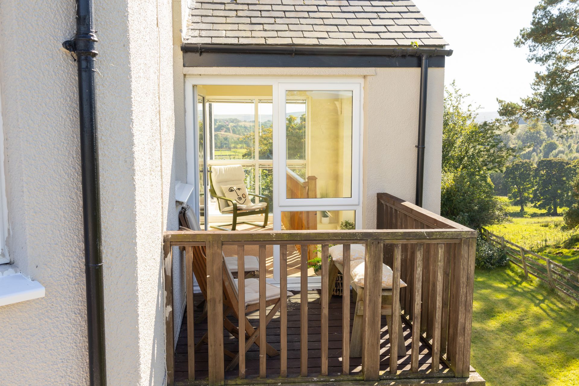 Small balcony with wooden railing outside a house, beside a glass door overlooking a sunny garden.