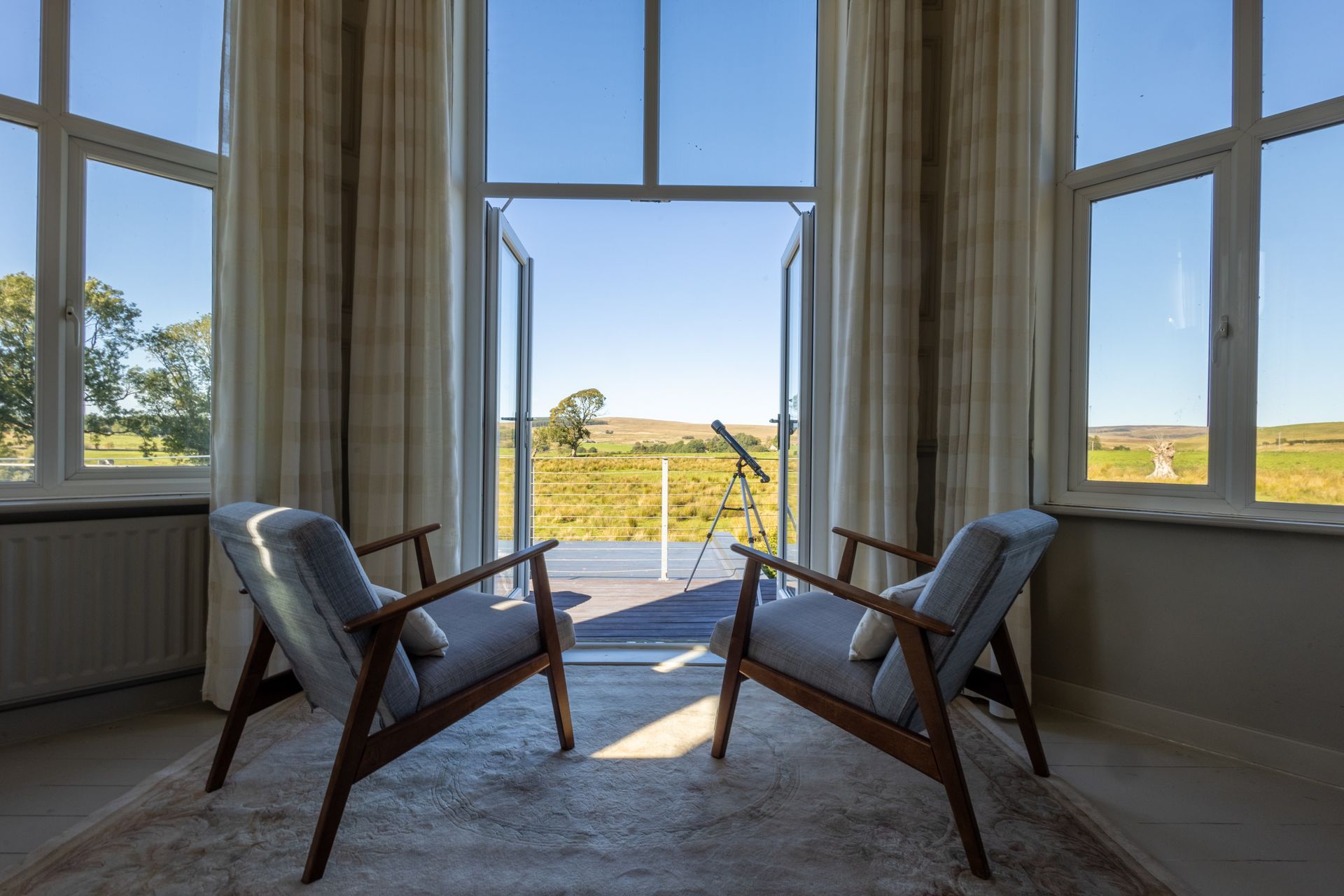 Sunlit room with two wooden chairs facing a large window overlooking fields