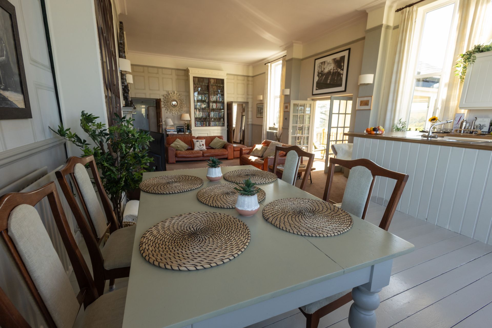 Bright dining room with a white table, woven placemats, wooden chairs, and sunlight from large windows.