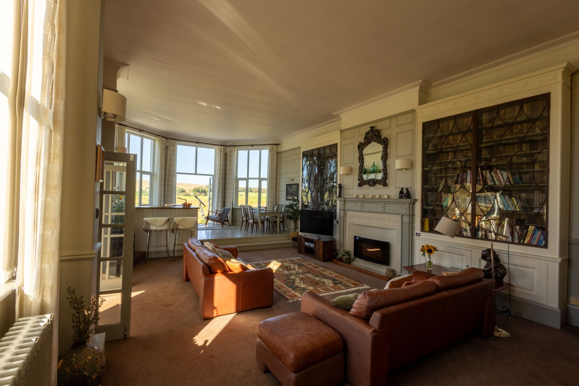 Sunlit living room with leather sofas, fireplace, bay windows, and built-in bookshelves