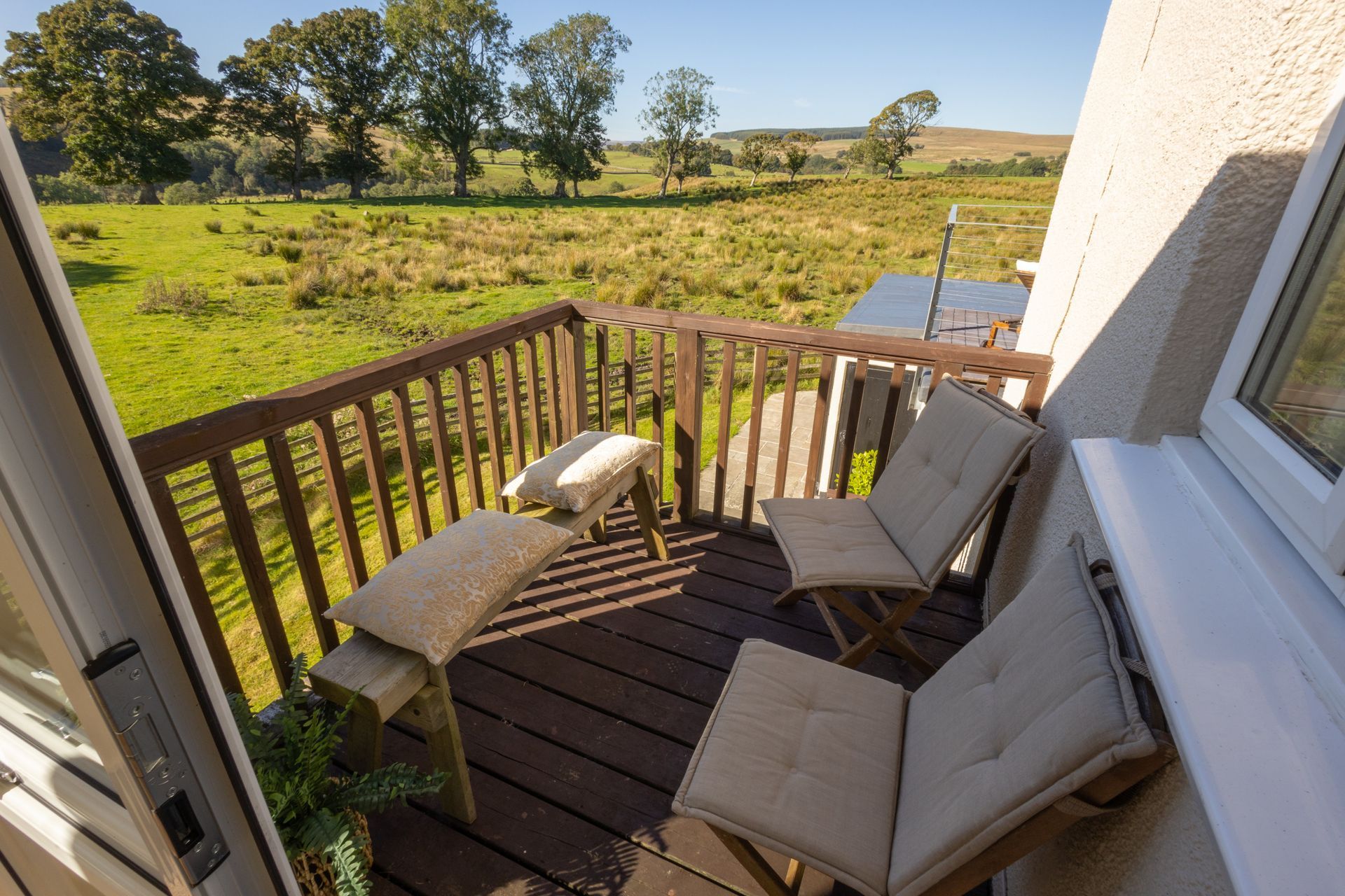 Small balcony with cushioned chairs and a bench overlooking a green field and trees.