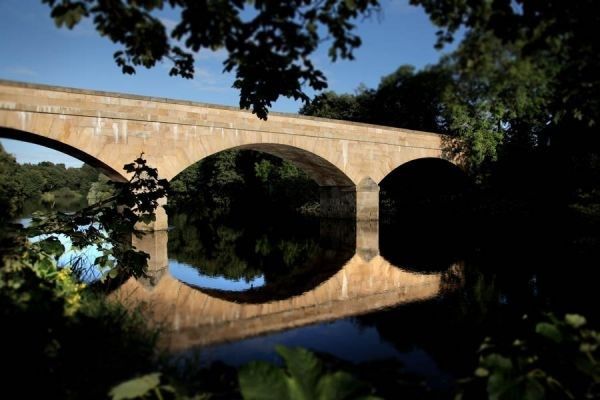 Stone bridge with three arches reflected in calm water, framed by trees and blue sky bellingham 