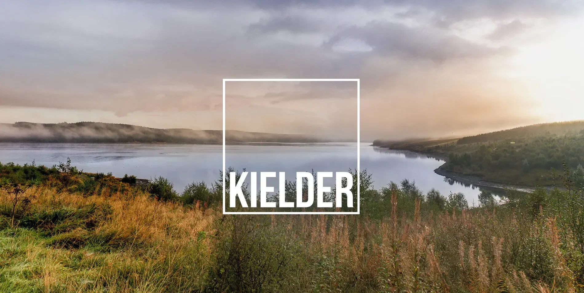 Kielder lake landscape at sunrise with misty water, hills, and autumn grass, framed by a white logo