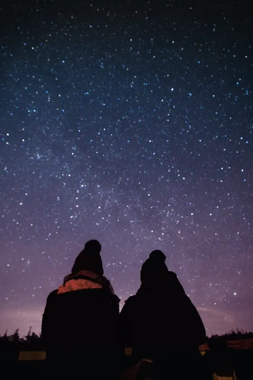 Two people in dark coats sit under a starry night sky with a purple glow near the horizon