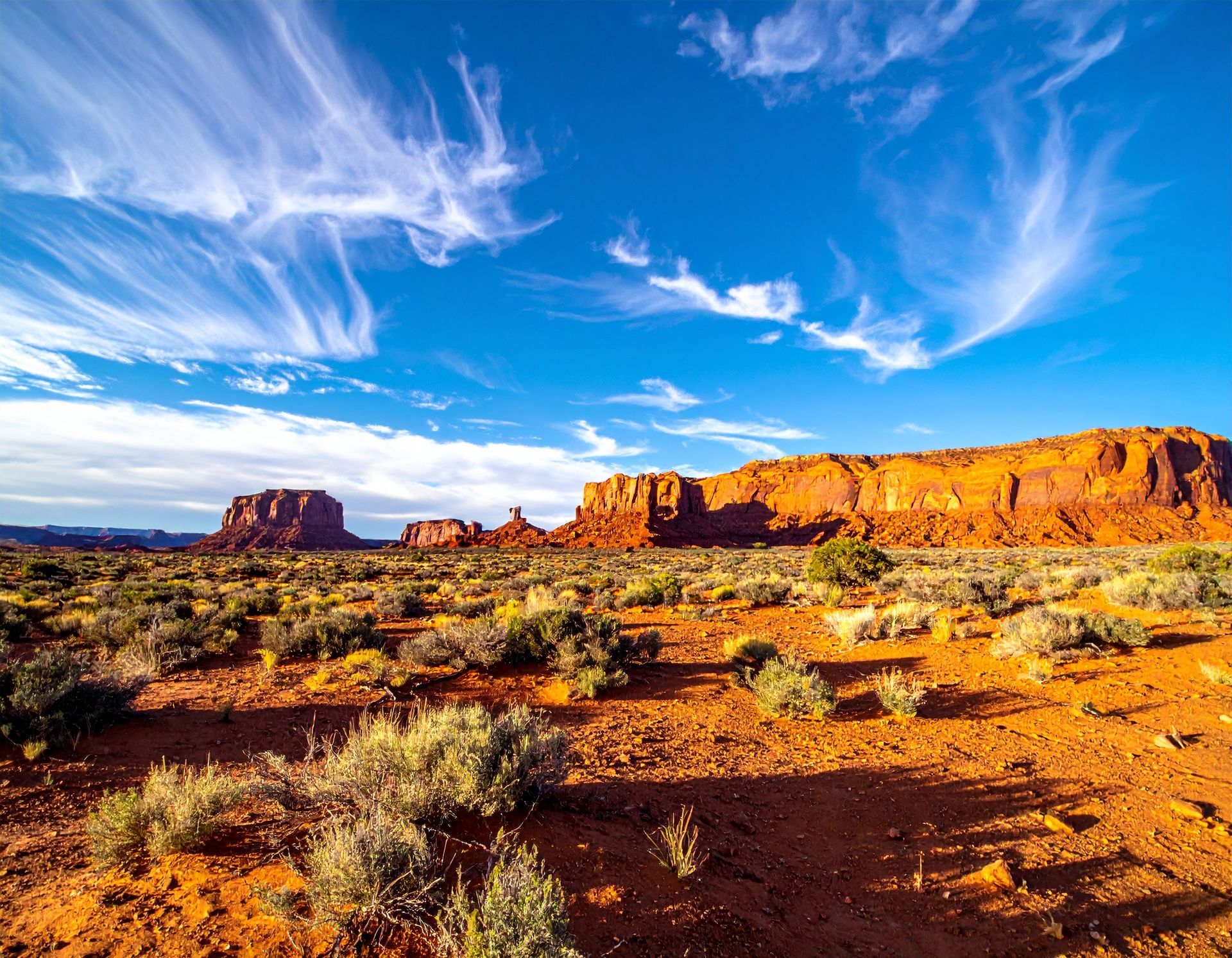 Sunlit red-rock desert mesa under a bright blue sky with wispy clouds.